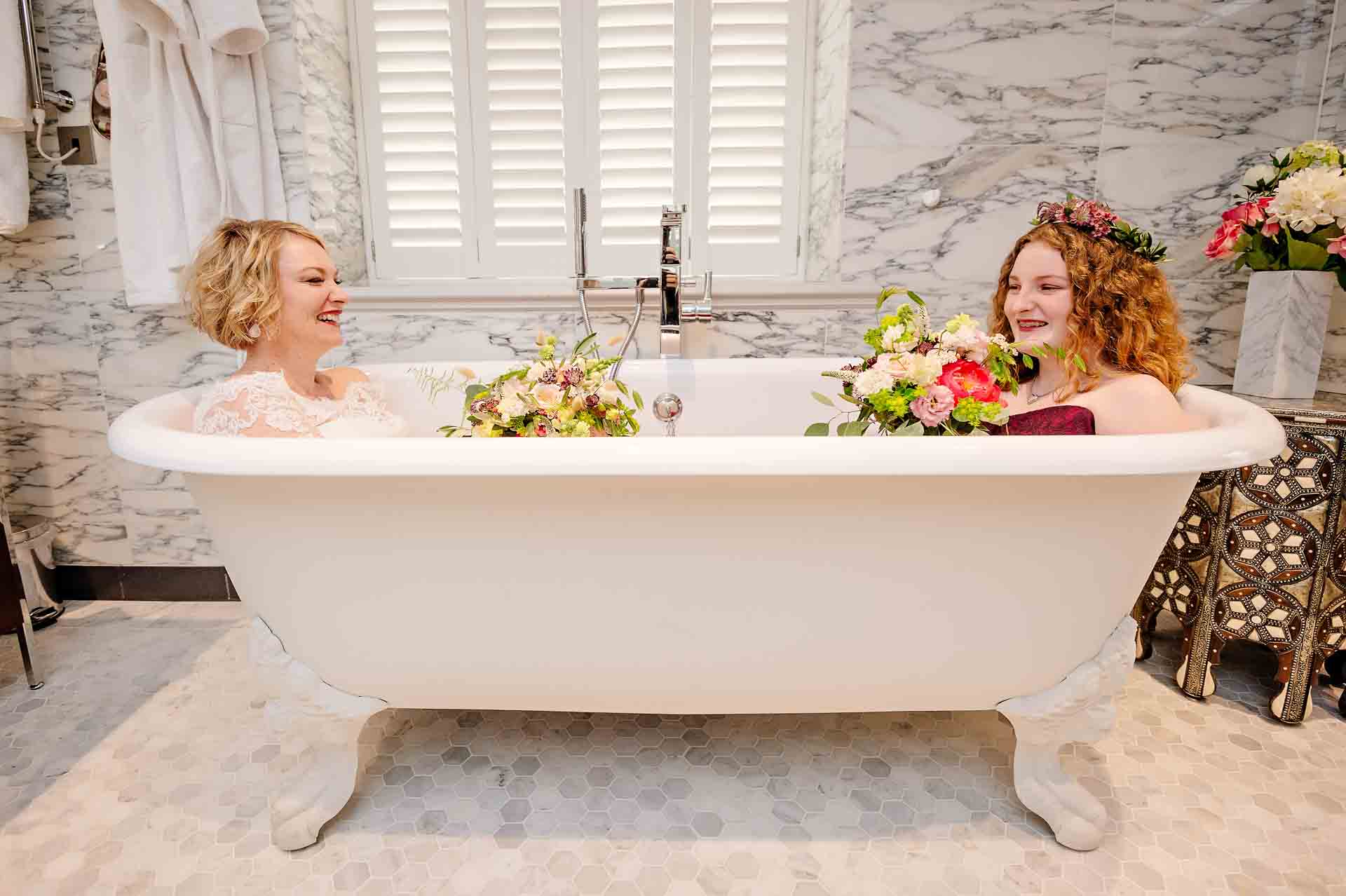 Bride with Bridesmaid laughing in bath during wedding preparations