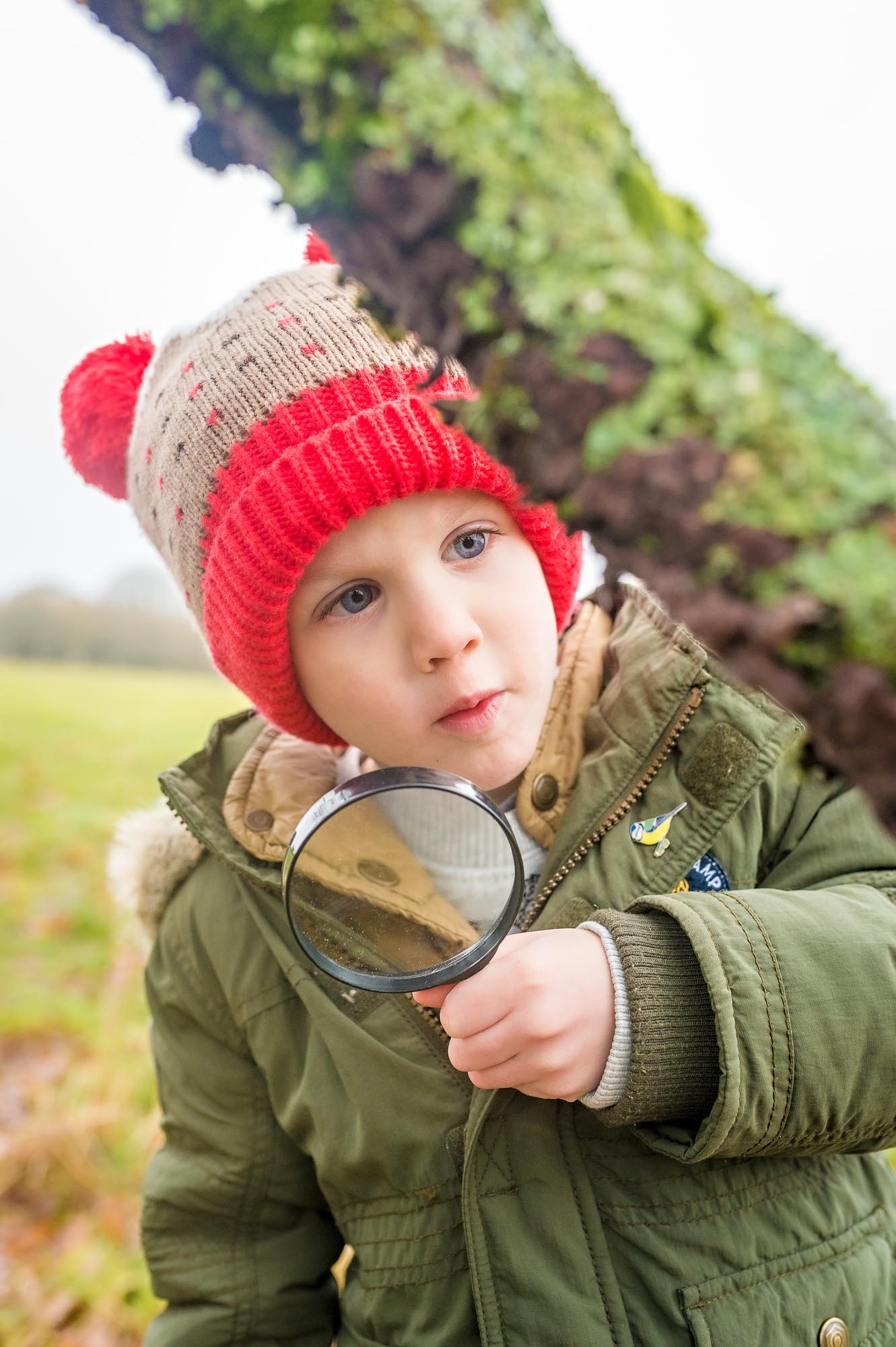 Little boy in bobble hat looking closely at branch holding magnifying glass