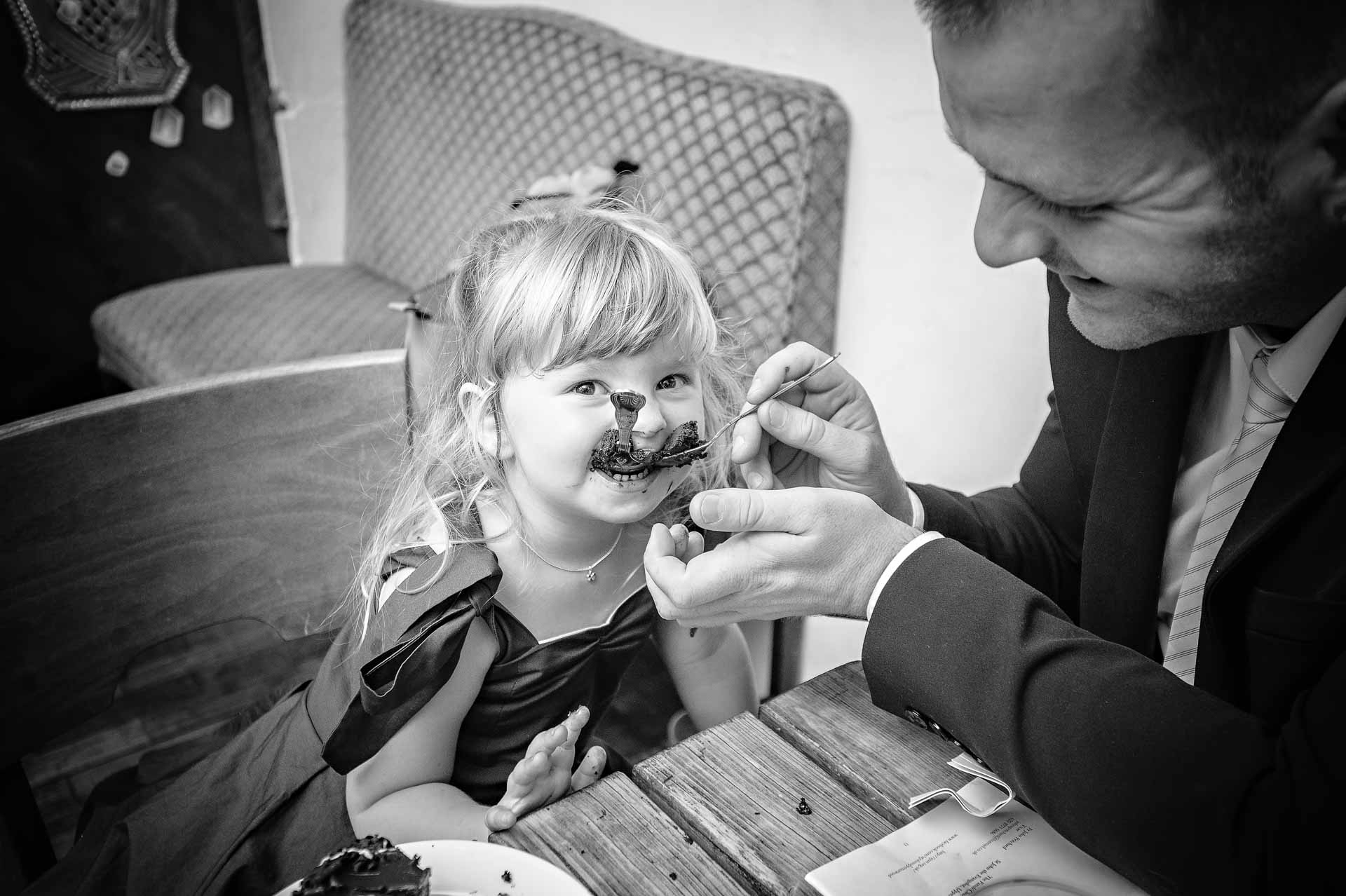 Little girl with two spoons in mouth being fed by dad at wedding meal