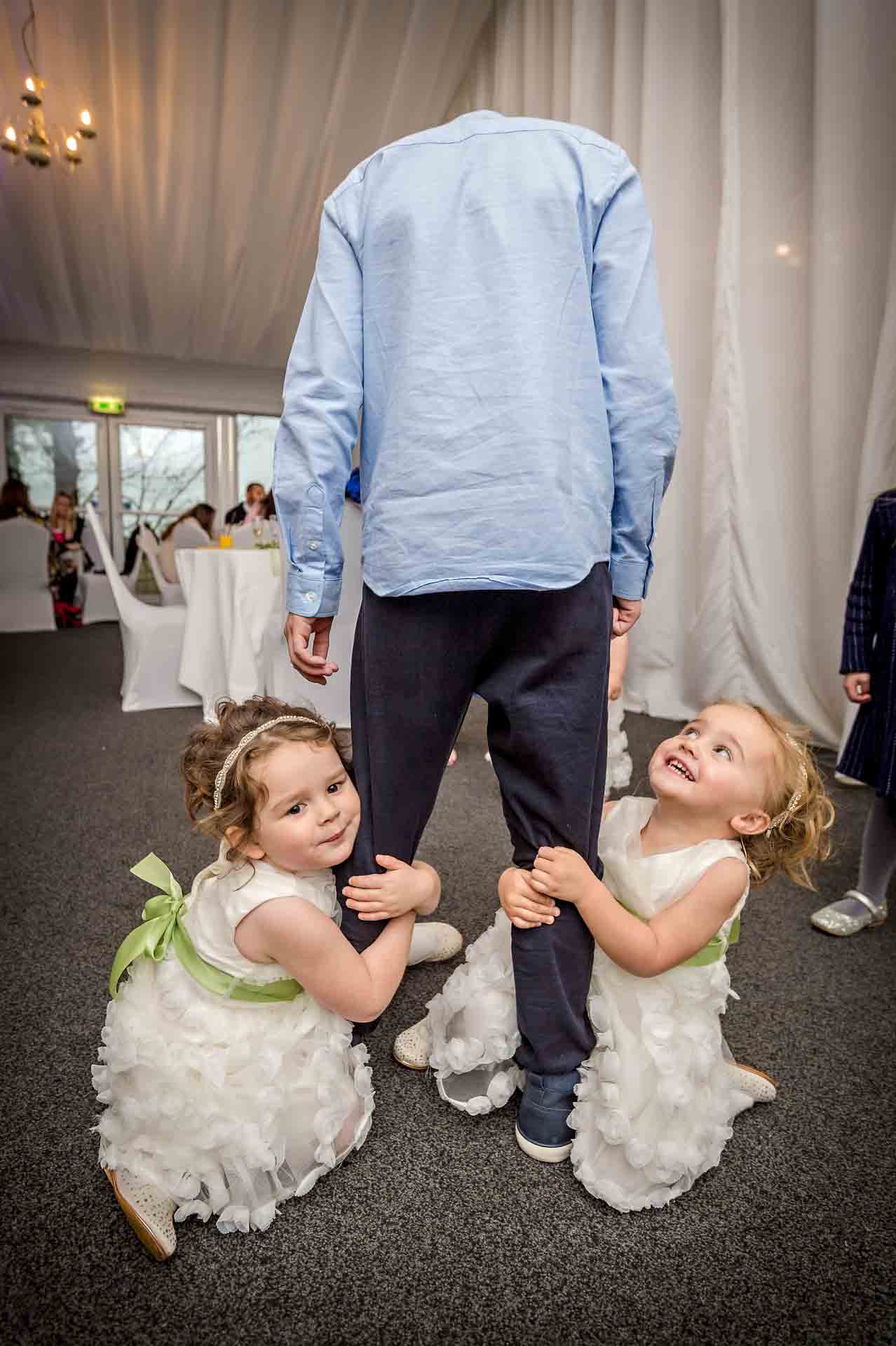 Two young bridesmaids cling on to leg of boy at wedding
