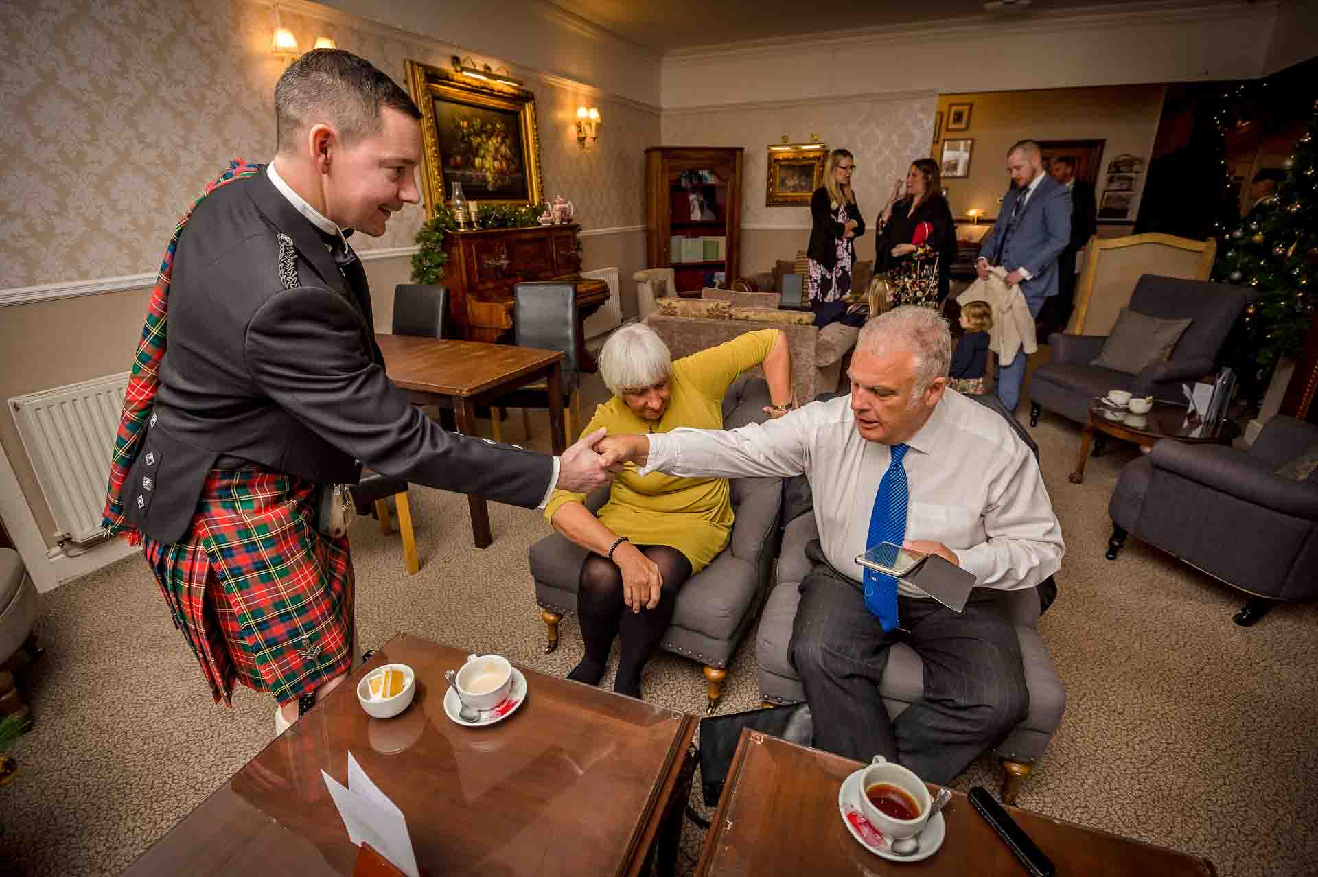 Groom in kilt shakes hands with guests in lounge at Llechwen Hall Hotel wedding