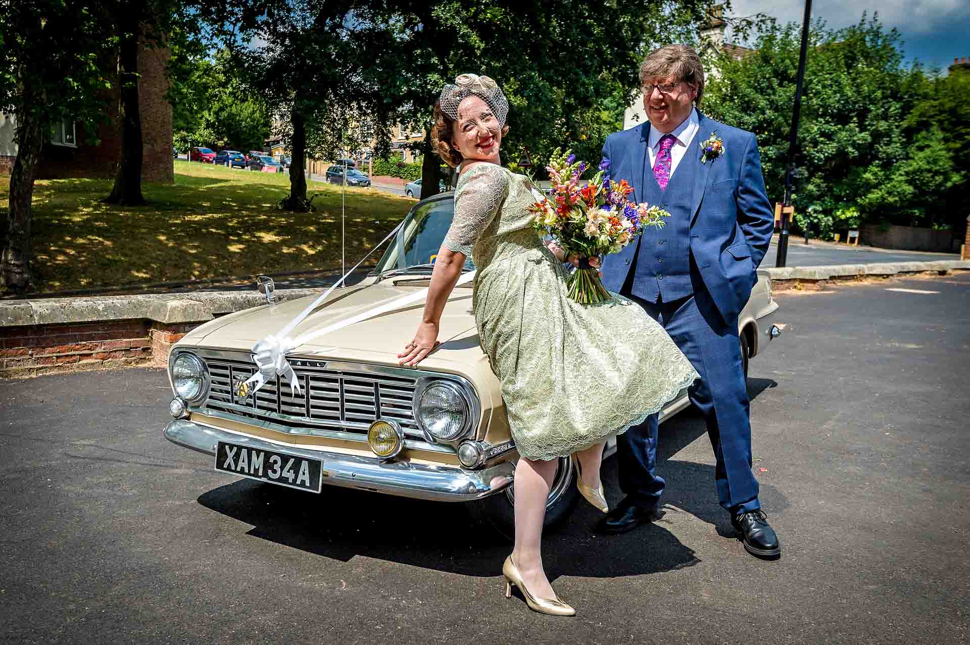Bride leaning on bonnet of vintage Vauxhall with father watching
