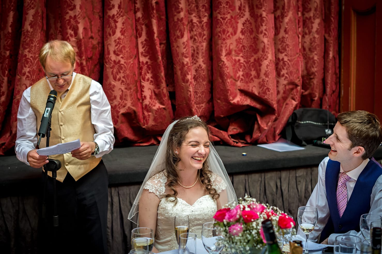 Bride laughing at father's wedding speech whilst groom watches