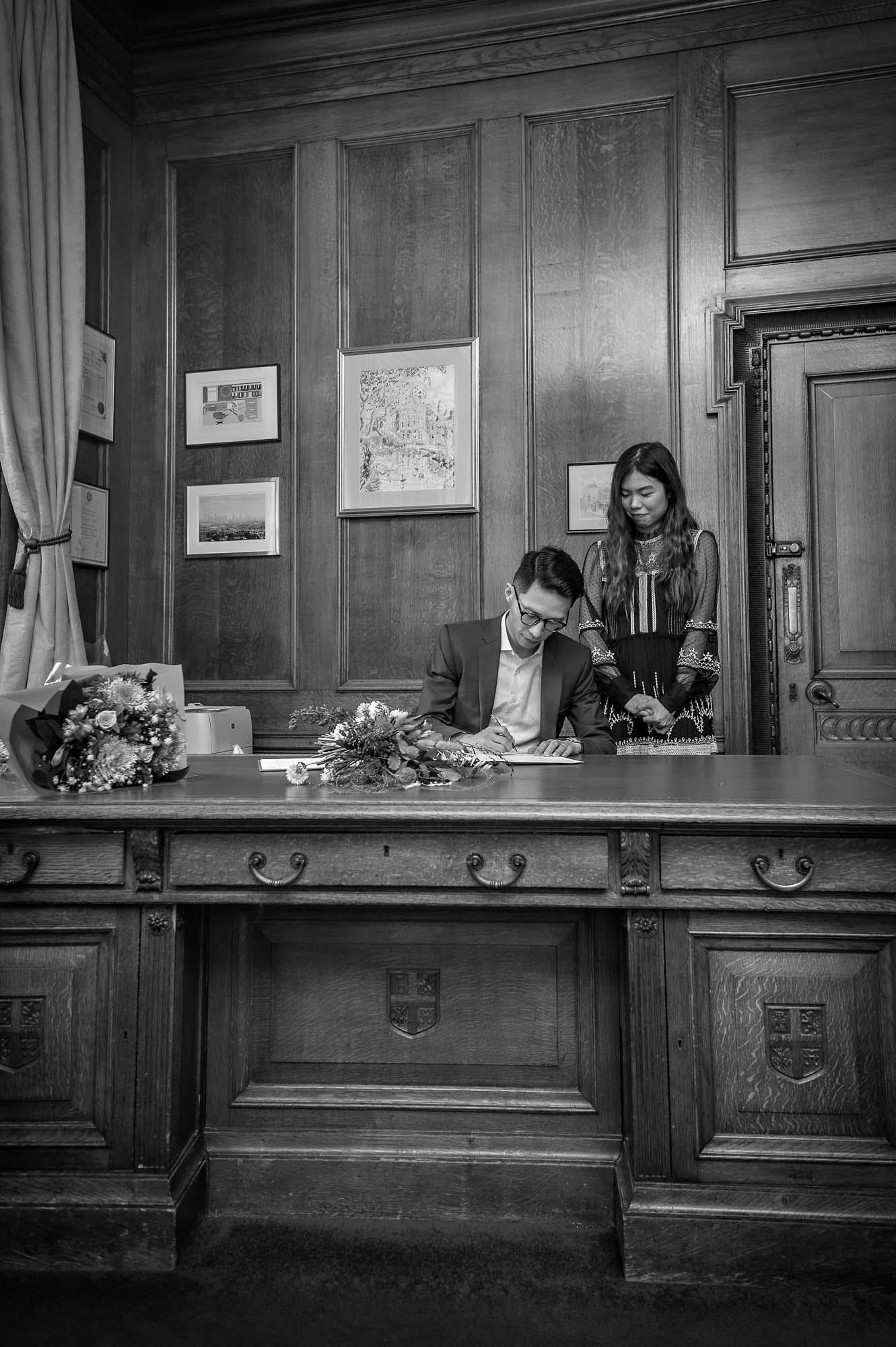 The groom signs the register whilst his bride watches on at Islington Town Hall