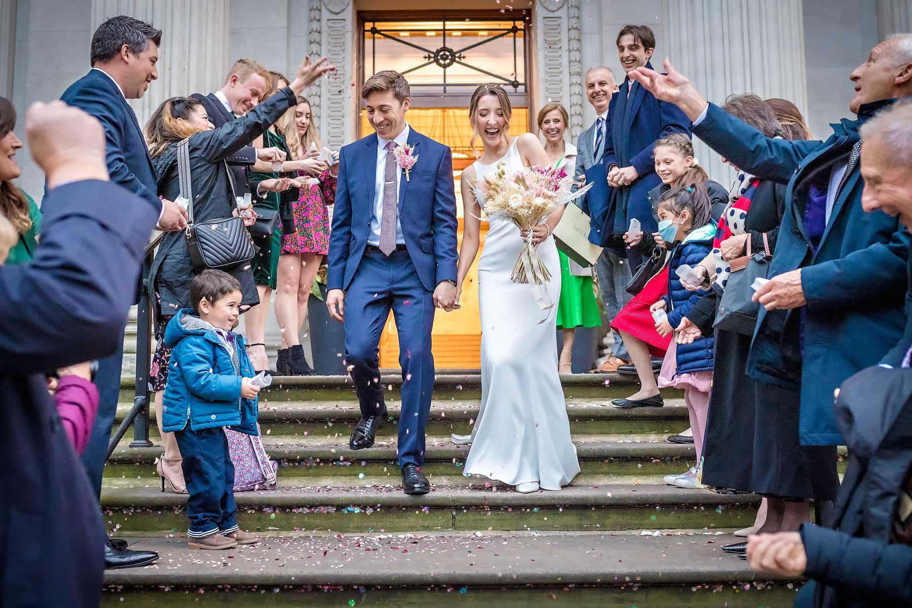 The laughing couple descend the steps outside Old Marylebone Town Hall whilst guests throw wedding confetti