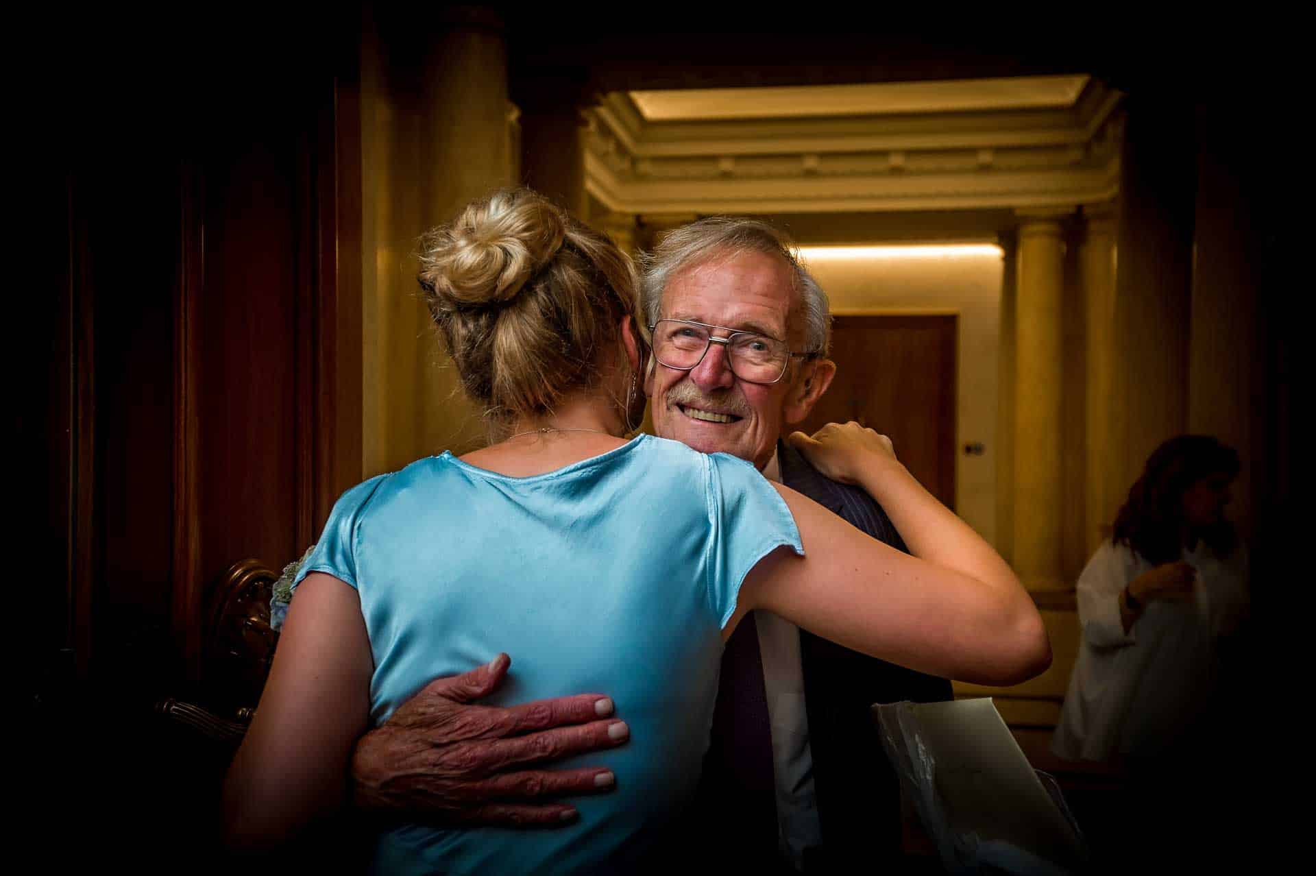 Bridesmaid with back to camera hugs elderly male relative who is smiling at the photographer