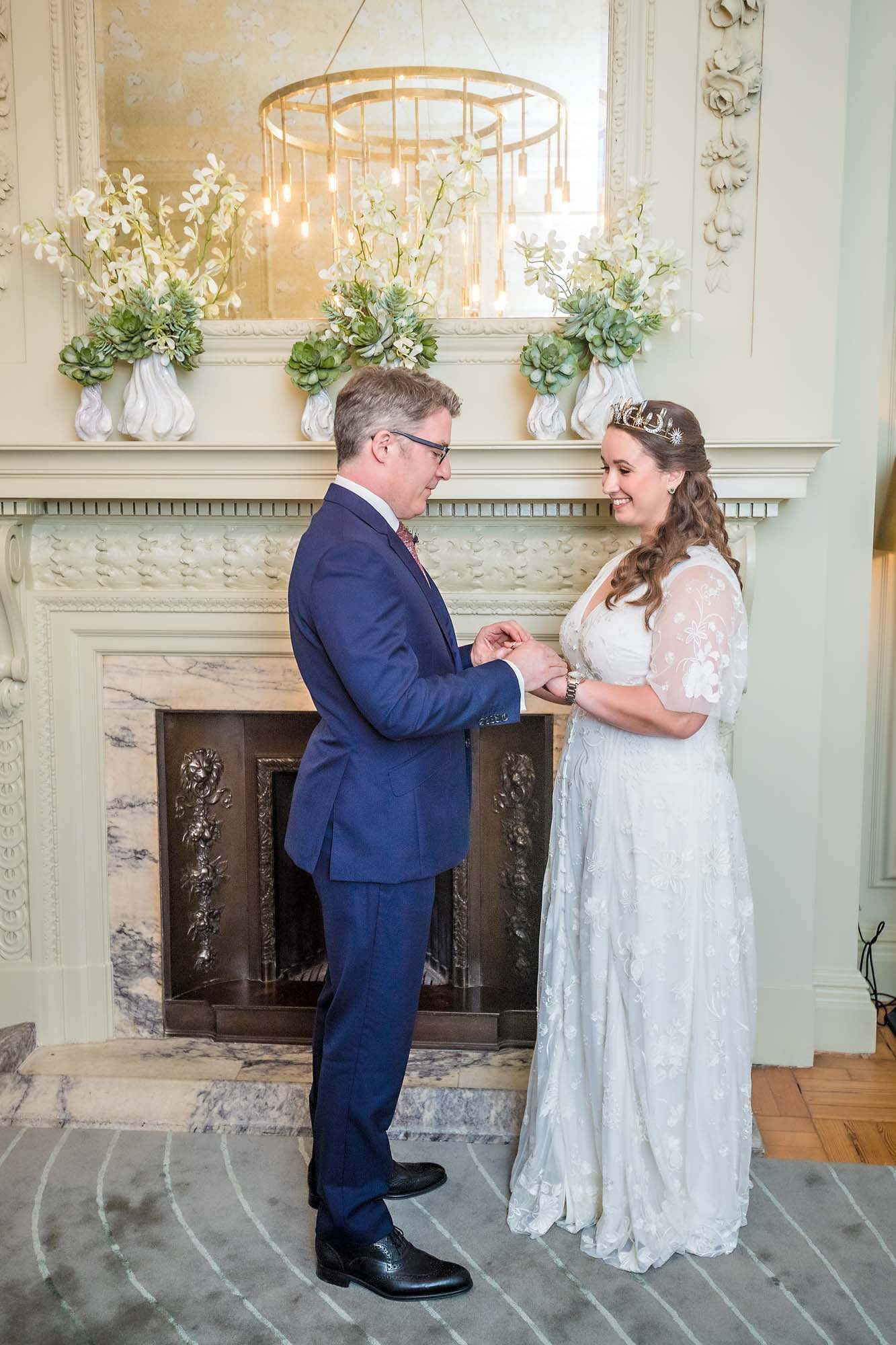 Groom placing ring on bride's finger in front of the fireplace in the Pimlico Room, Westminster