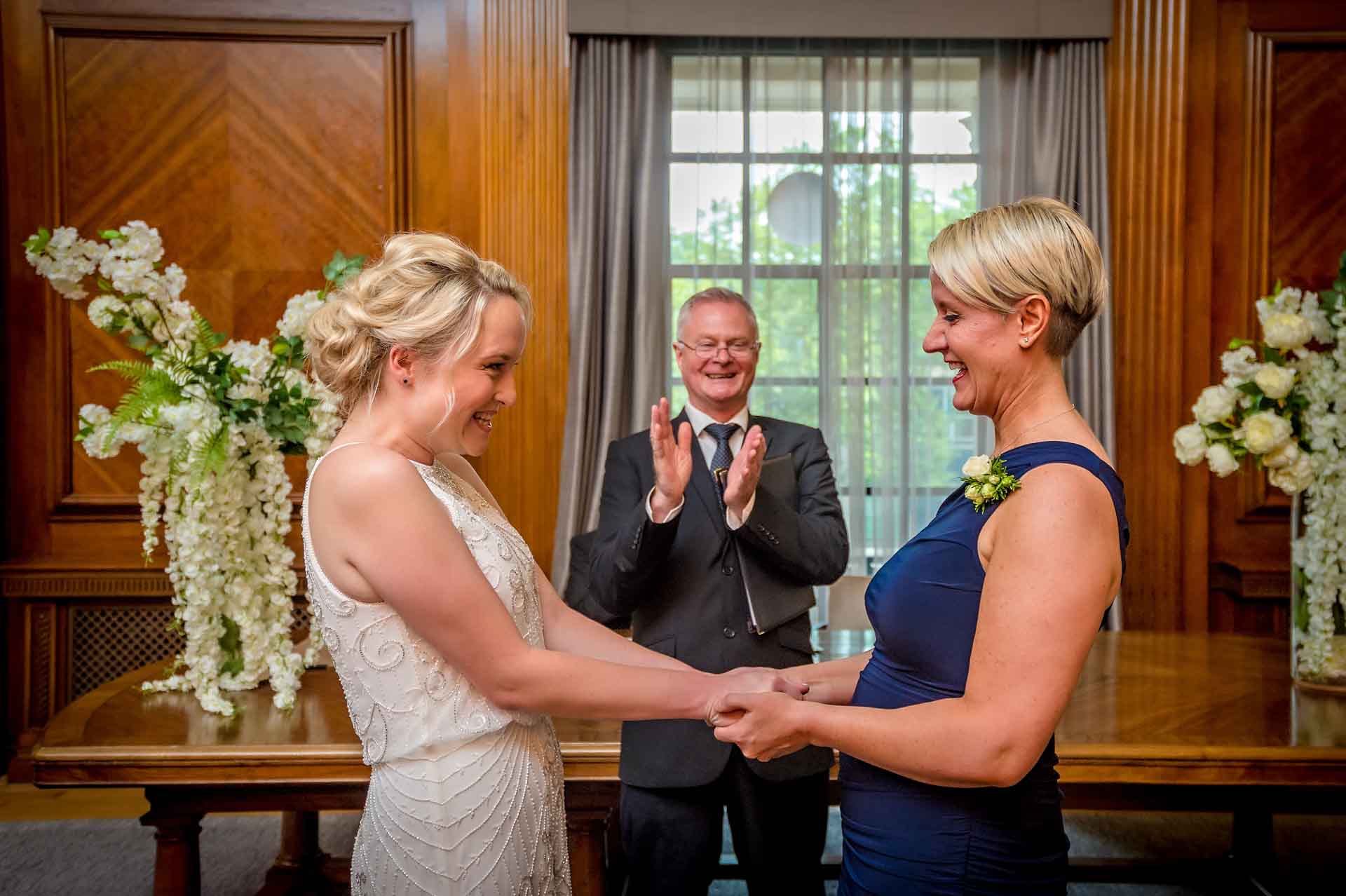Brides holding hands and laughing after LGBT marriage with registrar clapping