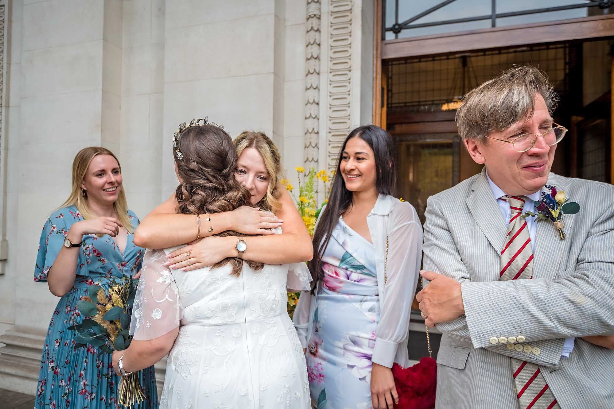 Female guests hugs the bride outside Old Marylebone Town Hall as other guests watch on