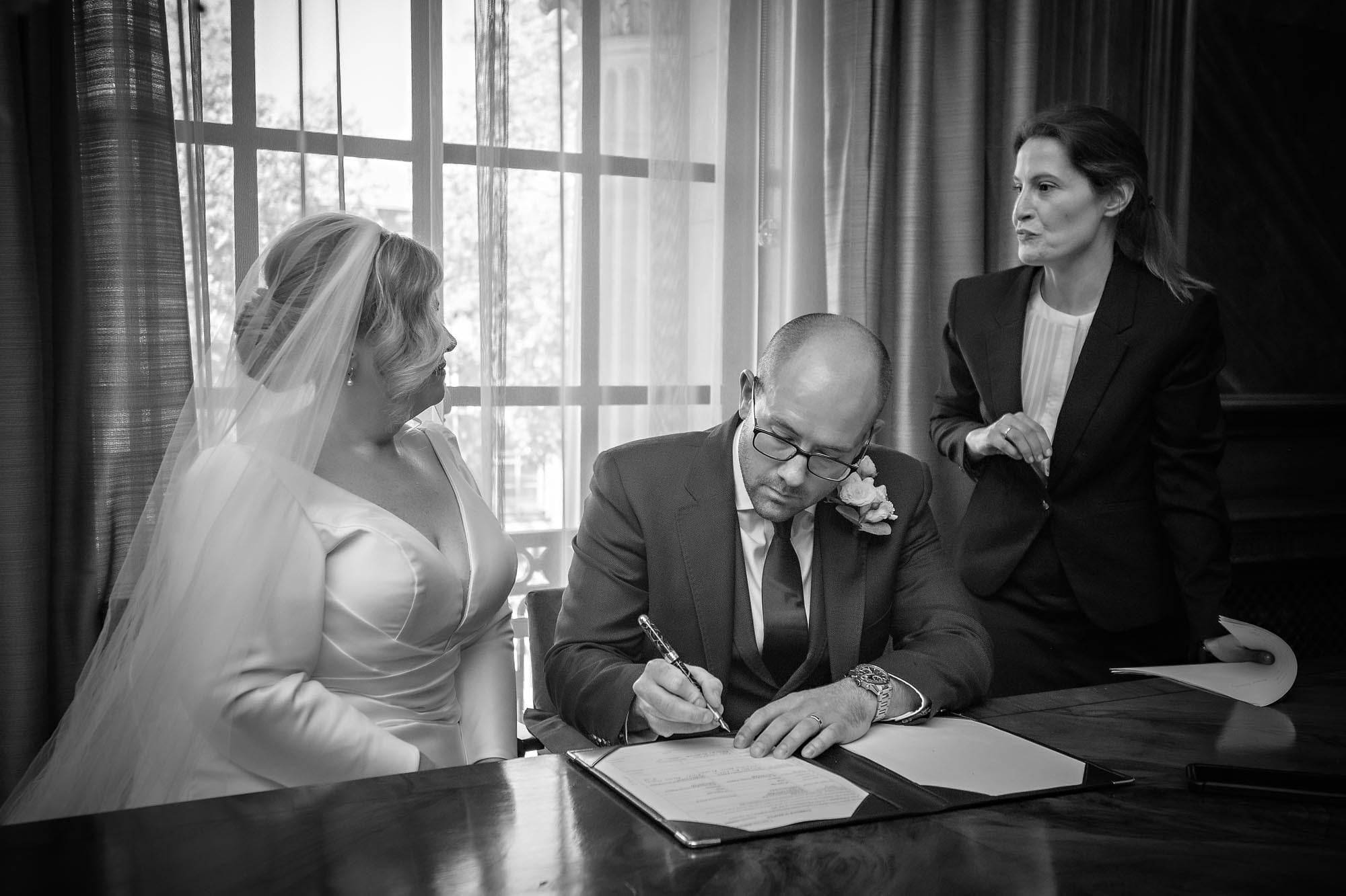 The groom seriously signs the regiaster the the bride and resistrar chat in Westminster Register Office