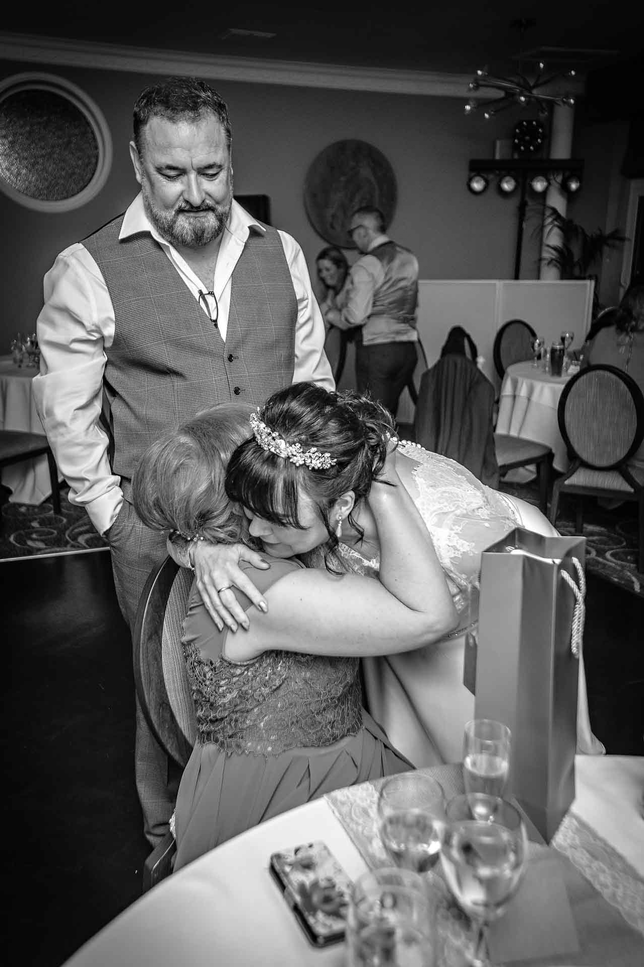 Black and White Photo of Bride Hugging Wedding Guest Whilst Groom Watches