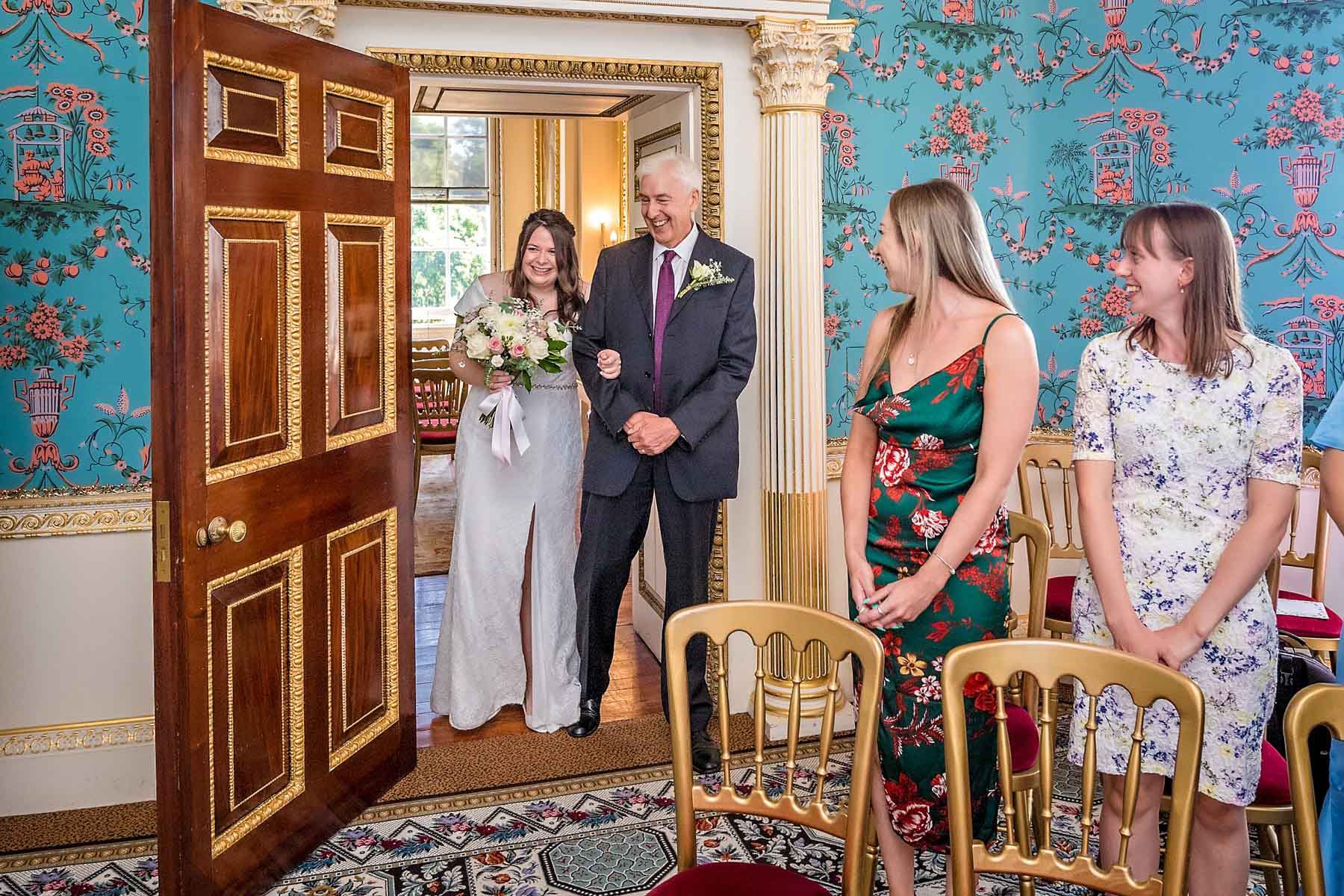 The bride enters the Salon at Danson House for her wedding ceremony. Walking with her father, she is peering in, looking for the groom as guests look on.