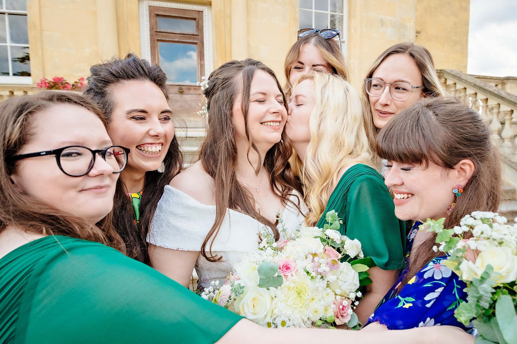 A bridesmaid kissing the bride on the cheek with friends gathered around