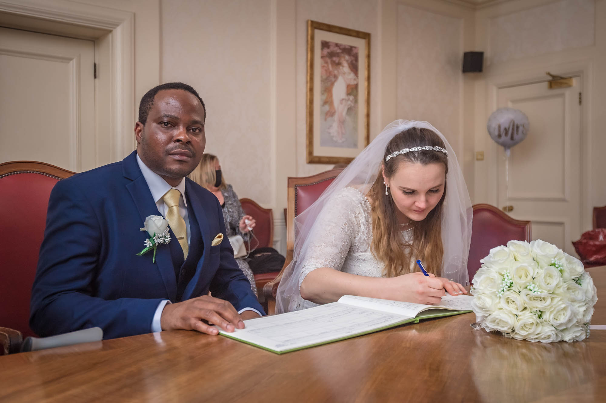 The bride signs the wedding register whilst a serious looking groom looks into the camera