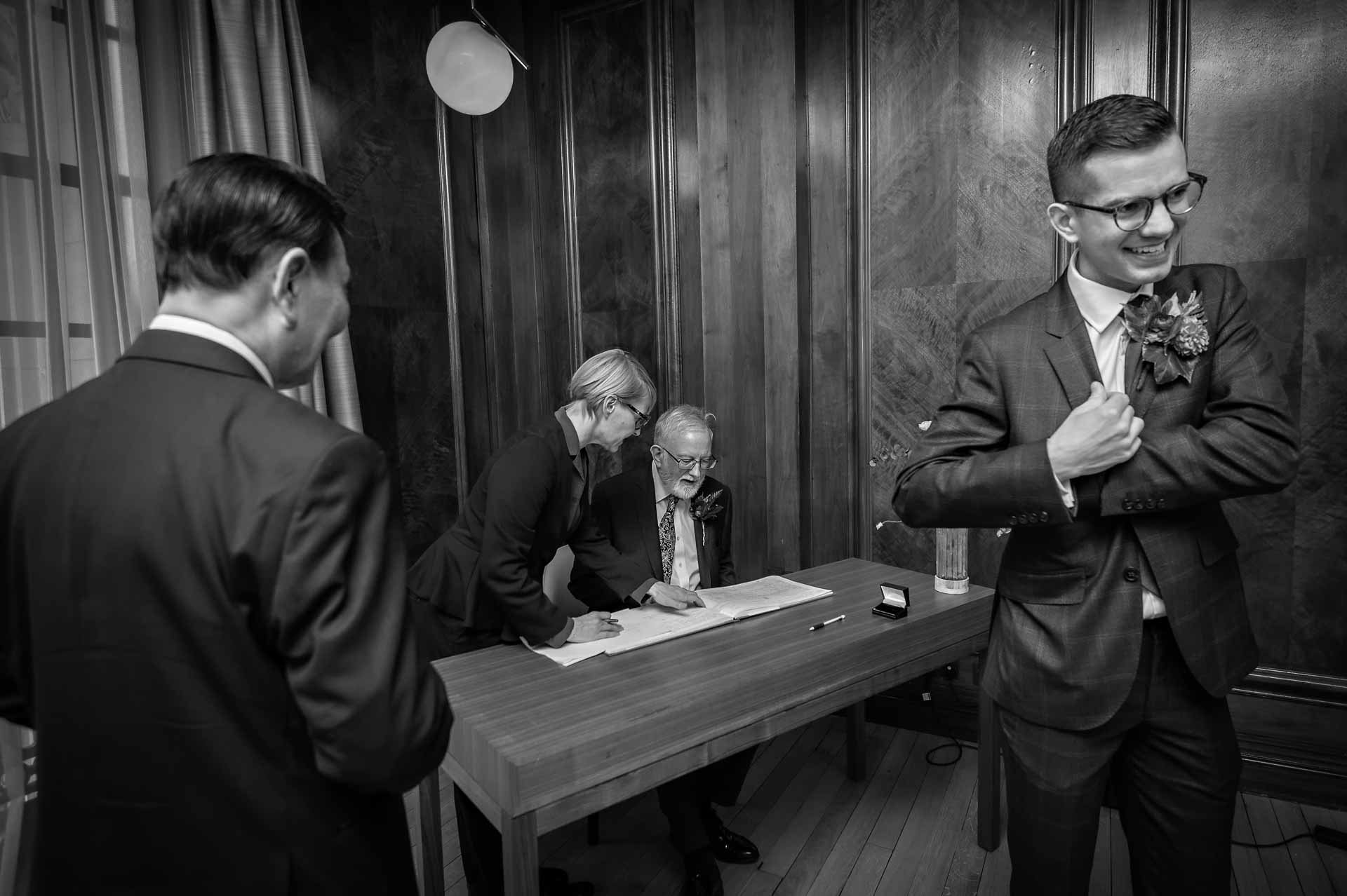 The groom pockets the marriage certificate as his father signs the register in the Marylebone Room, Westminster