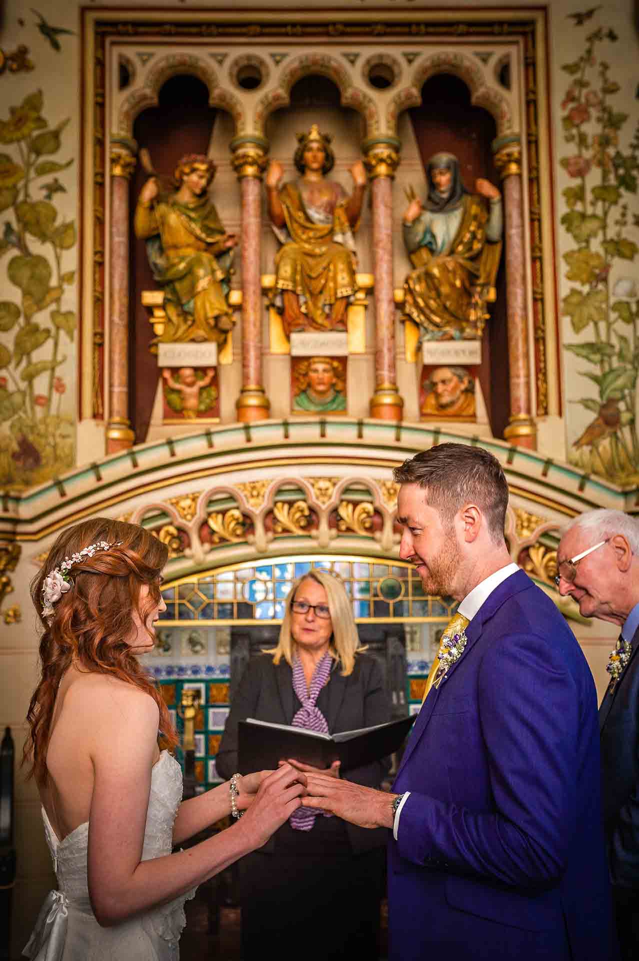 Bride places wedding ring on groom's finger at Castell Coch with three fates in background