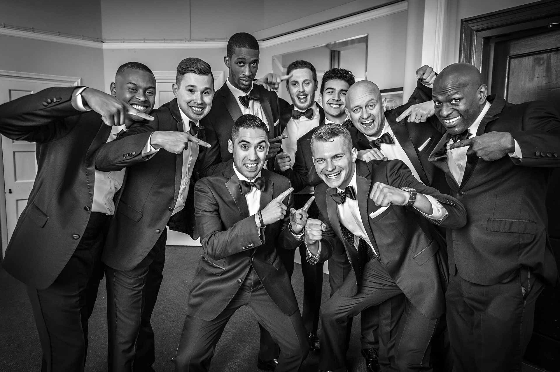 8 Black Tuxedo Dressed Groomsmen Pointing at Groom at Newport Wedding