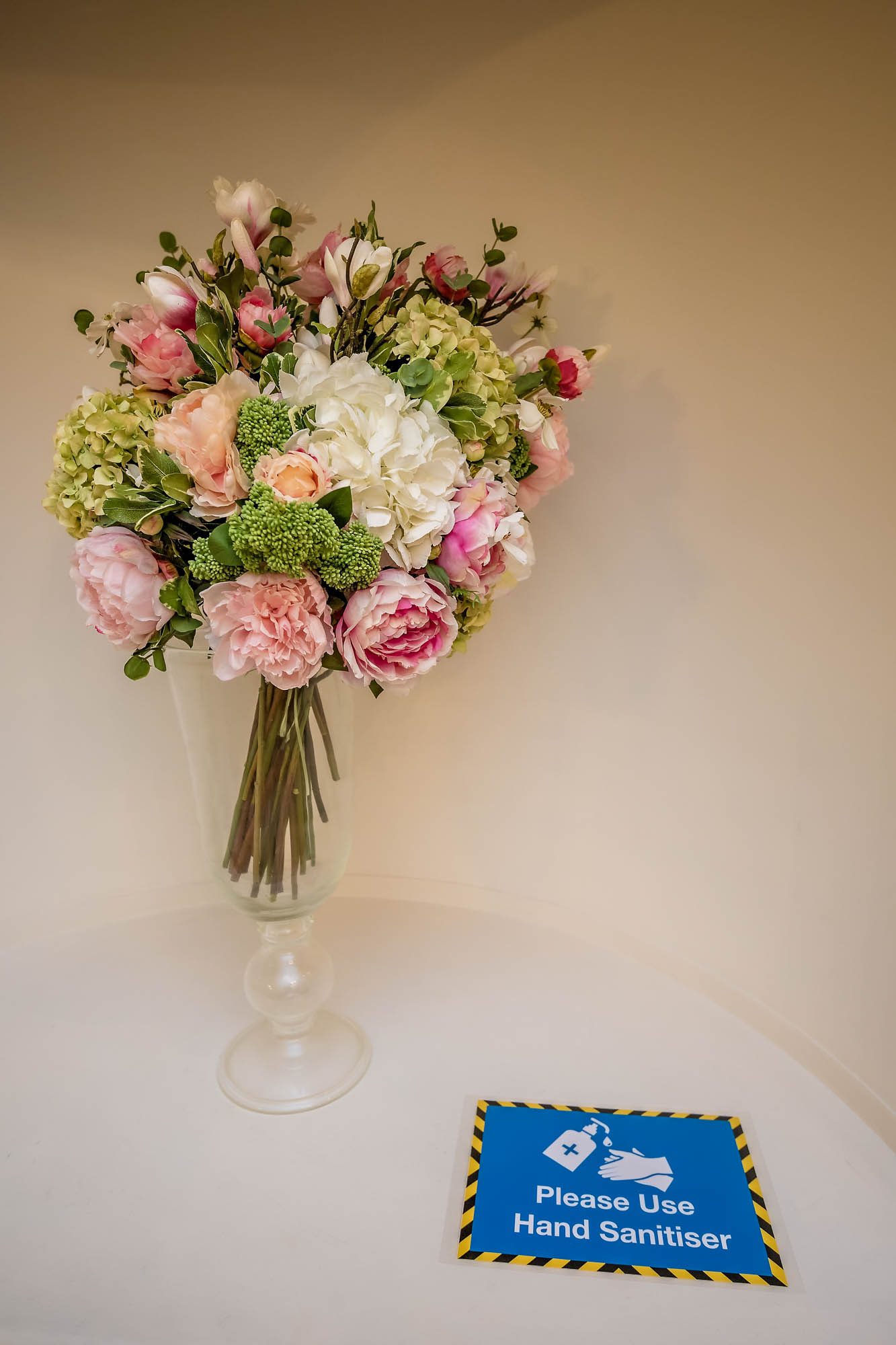Wedding bouquet in glass vase with blue 'Please Use Hand Sanitiser' sign.