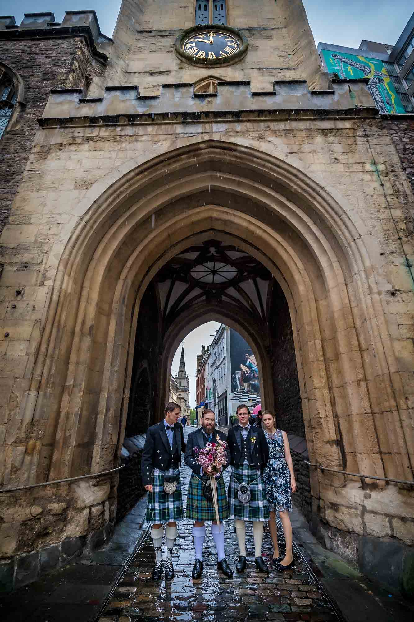 The kilted groom and best man and others under St John on the Wall's Church Clock Tower Arch in Bristol