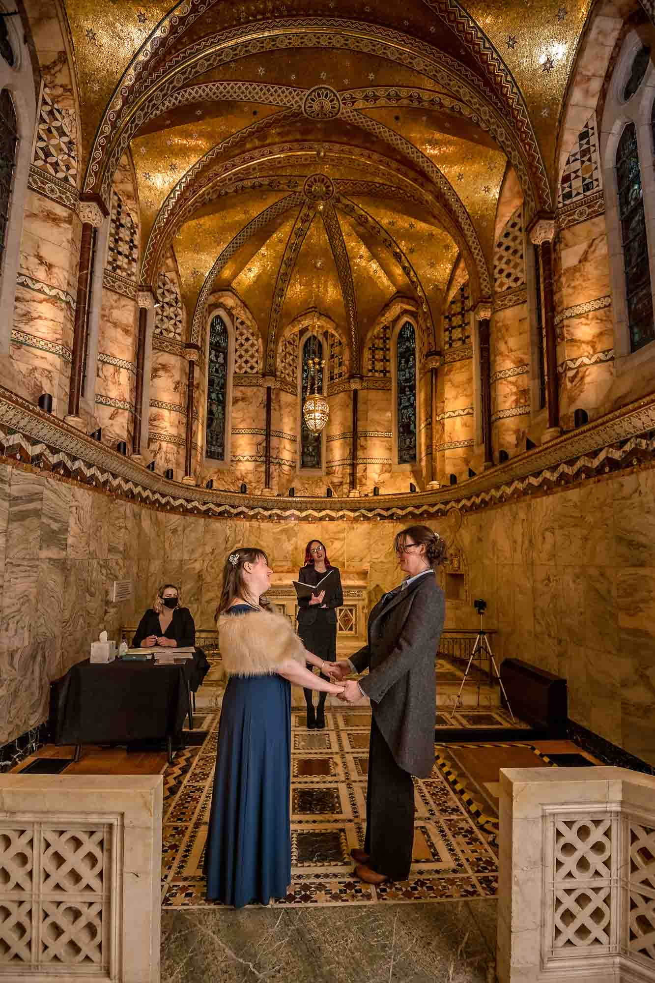 Female couple holding hands at their Fitzrovia Chapel wedding ceremony showing ornate ceiling