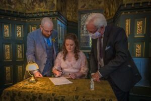 Bride signs the marriage schedule whilst groom and masked registrar watch on at Castell Coch, Cardiff