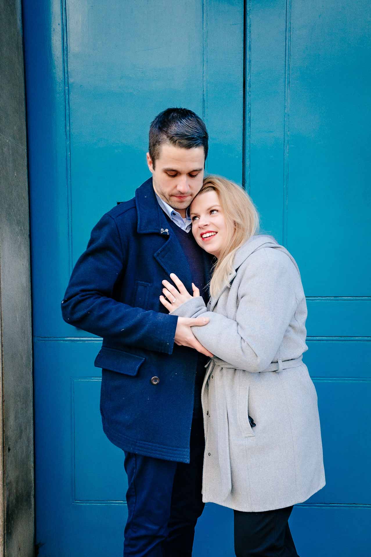 Newly-engaged couple in front of blue doorway with woman looking very happy