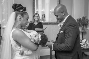 The groom places a ring onto his bride's finger as their baby that she's holding stares at the camera