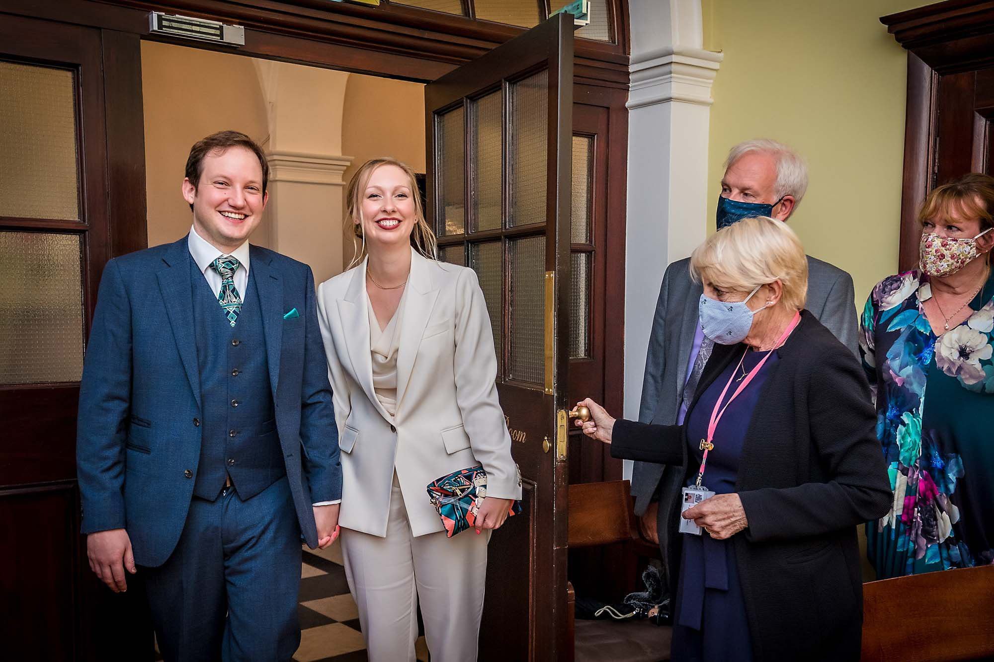 Smiling couple enter the Rossetti Room at Chelsea Old Town Hall with masked guests watching during Coronavirus pandemic