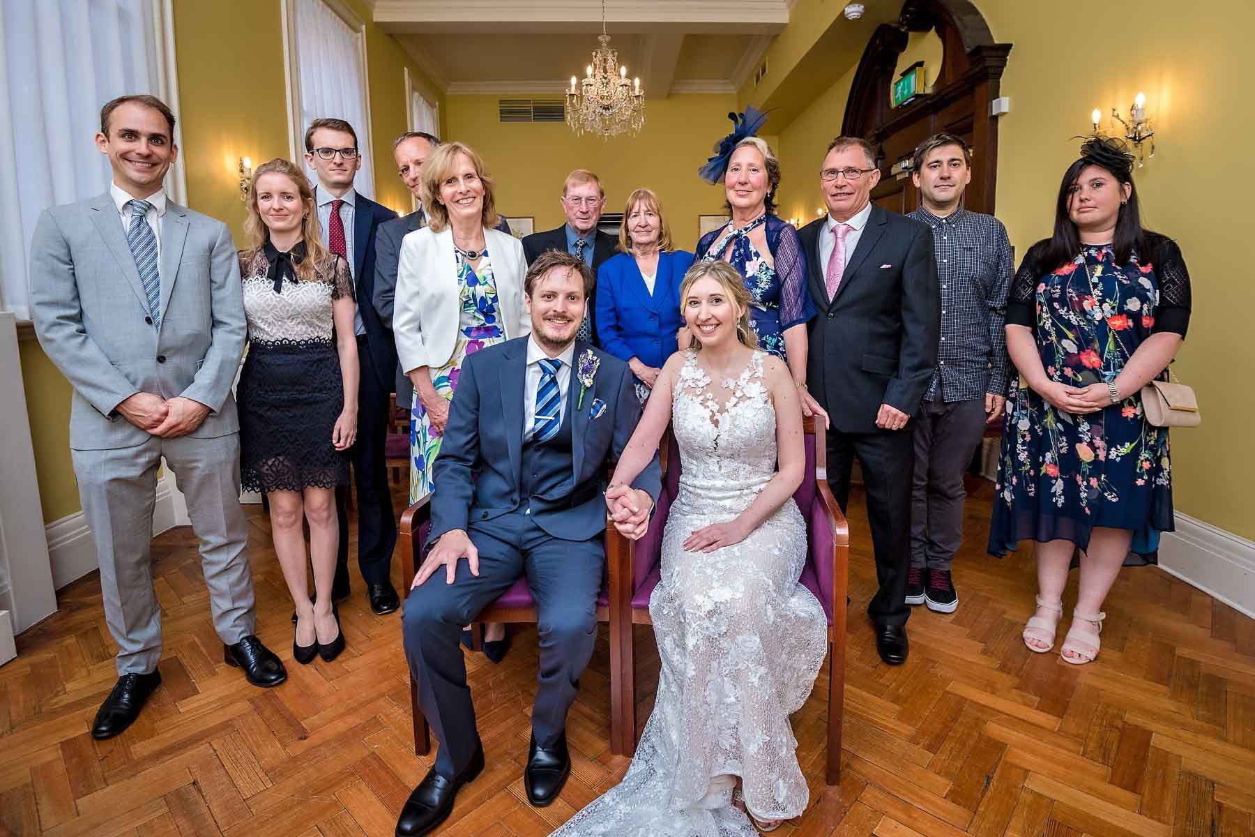 The seated bride and groom hold hands with entire wedding party standing behind them in the Brydon Room in Chelsea