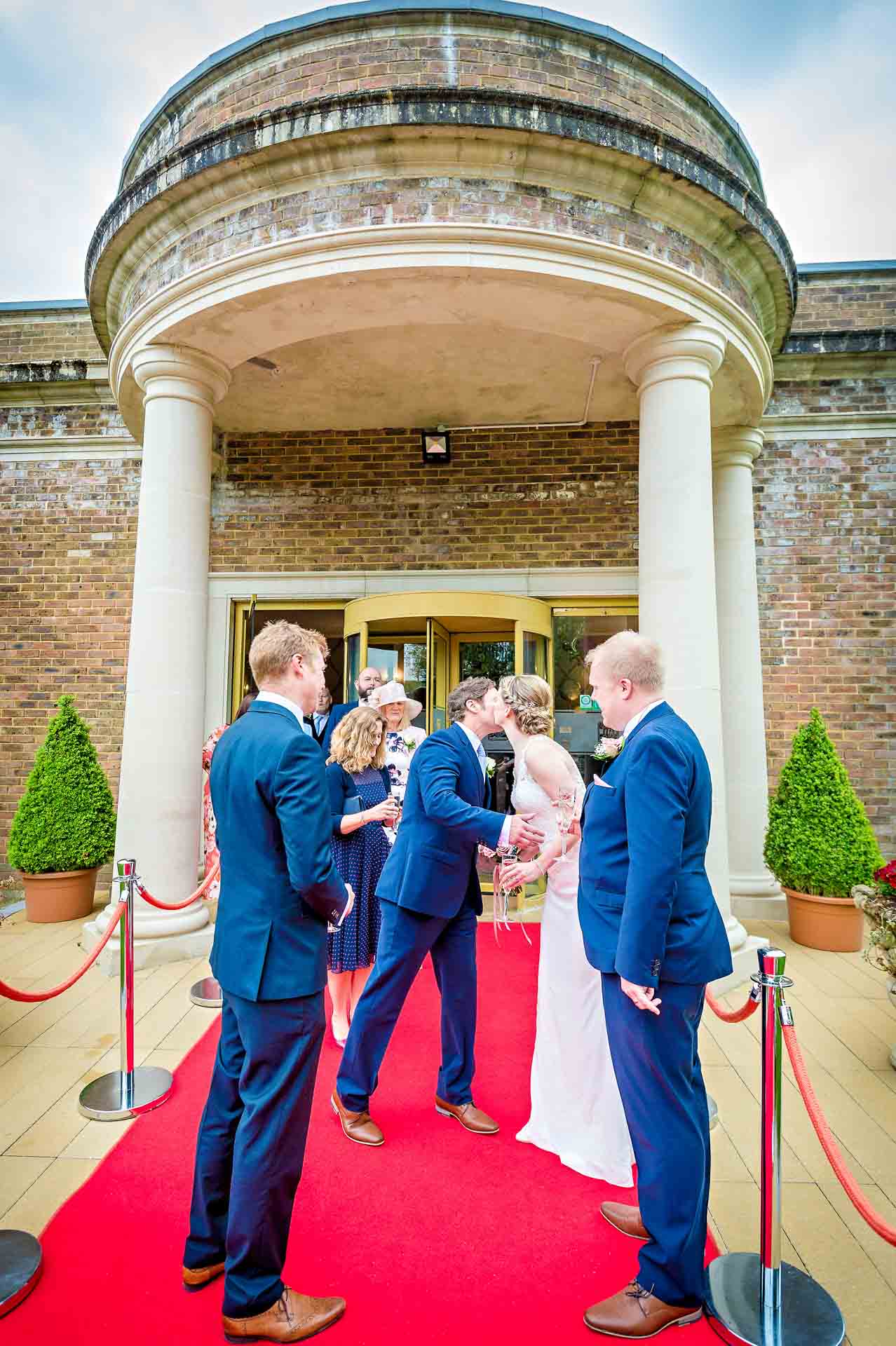 Bride kisses male guest at wedding whilst groom watches outside DeCourceys Manor