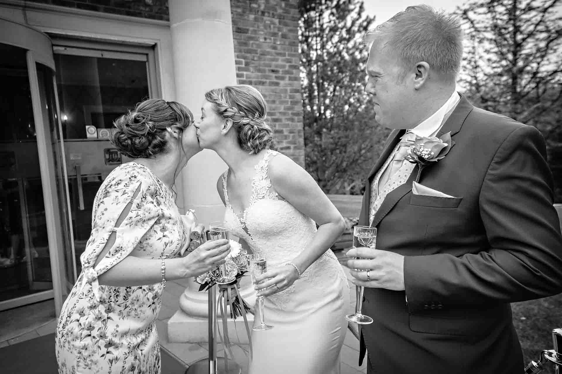 Bride kisses female guest at wedding whilst groom watches