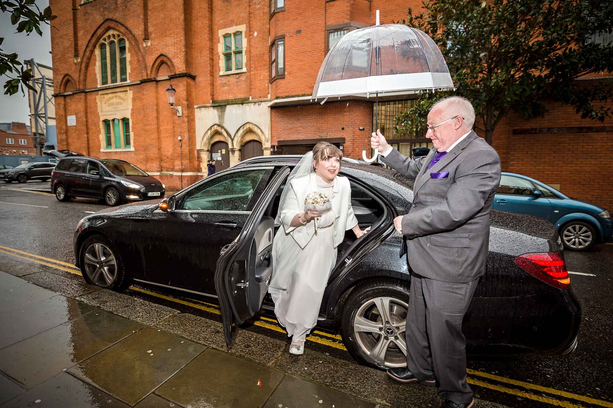 Bride leaving taxi in rain outside Chelsea Old Town Hall with father holding unbrella
