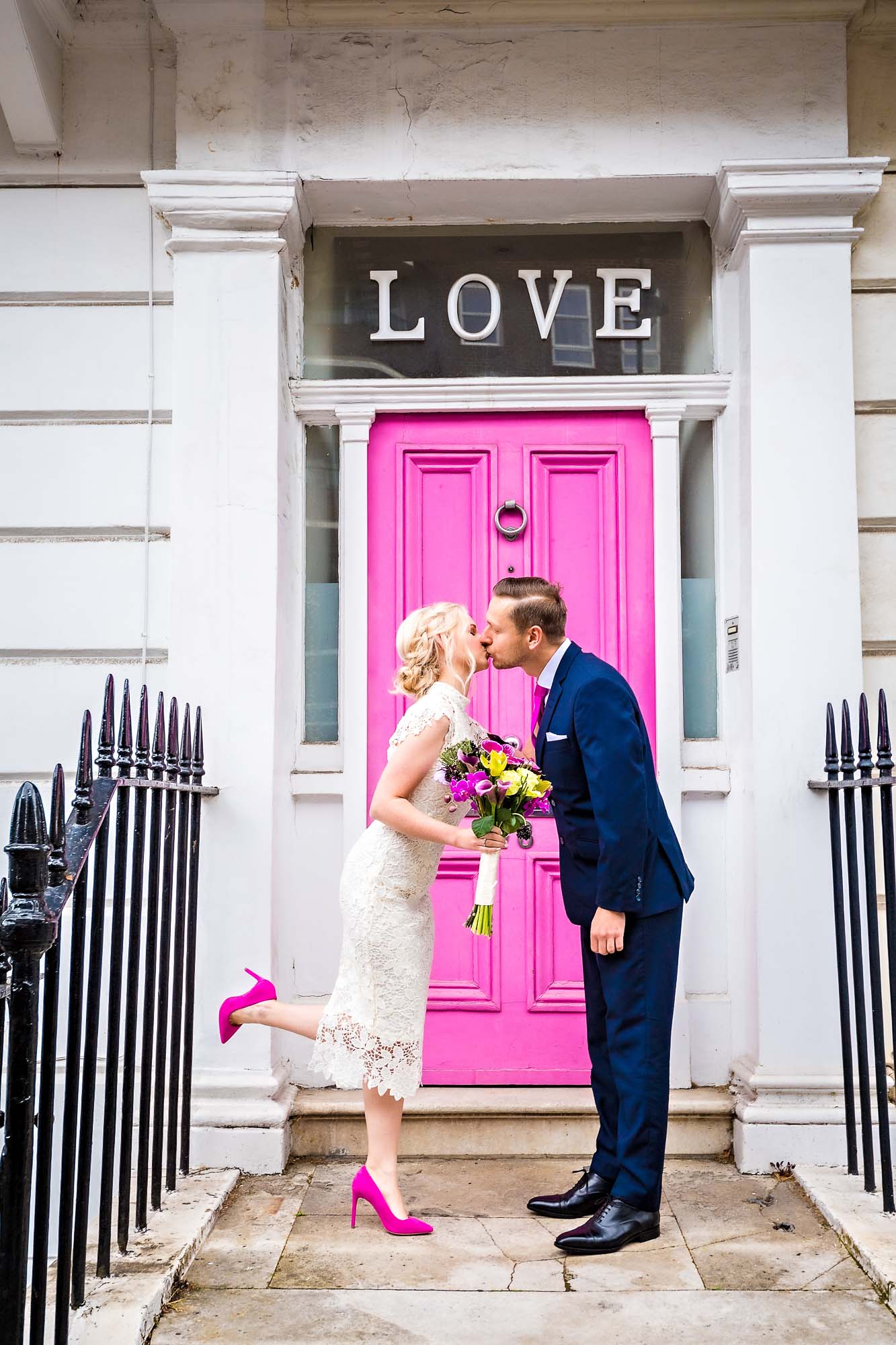 Bride in pink shoes kisses smart groom in front of pink door with 'Love' written above it