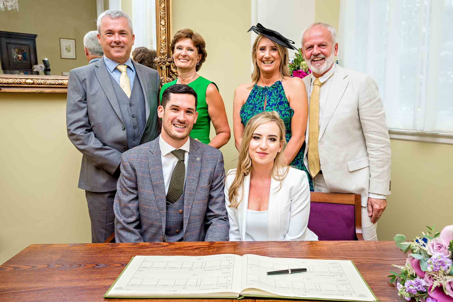 Bride and Groom with both sets of parents behind register signing table in Brydon room, Chelsea