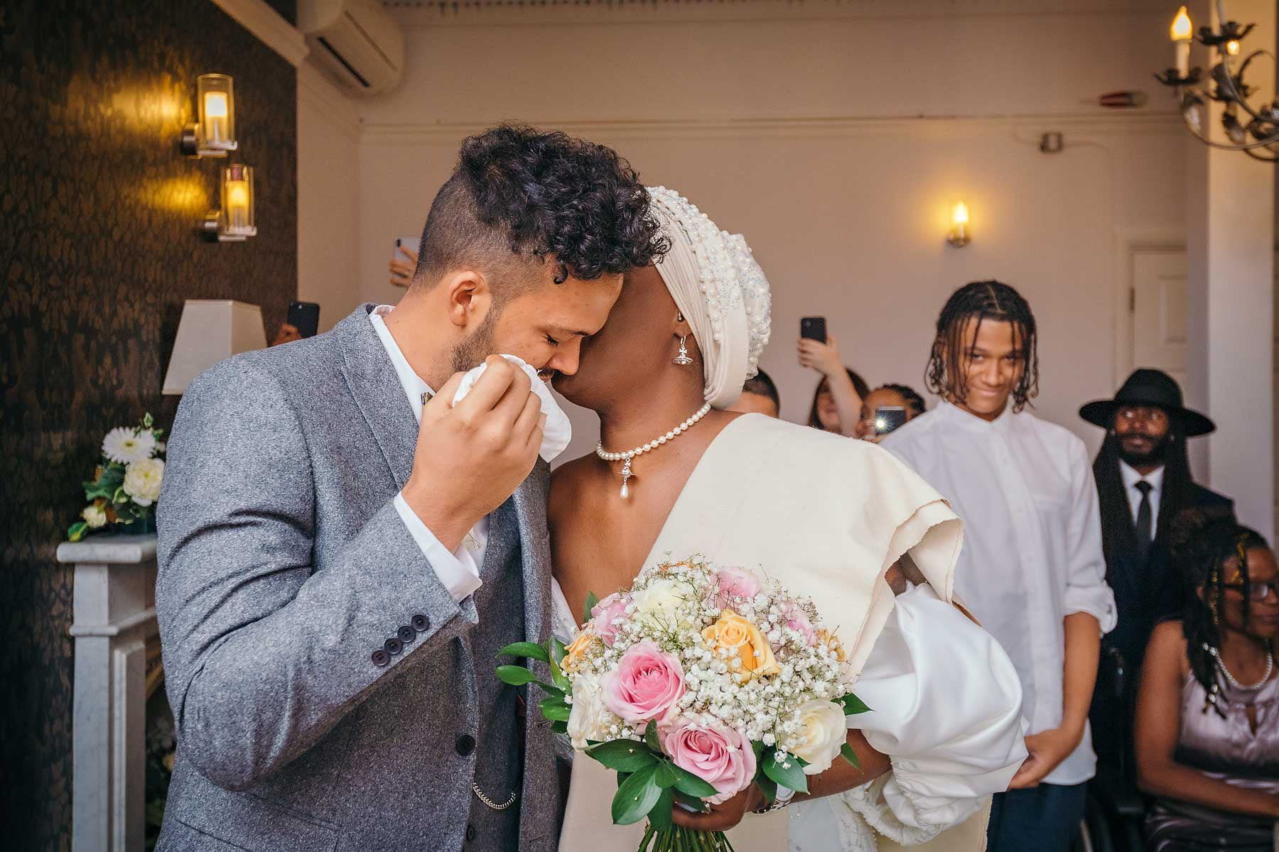 The bride kisses her groom as her wipes away a tear at a London wedding