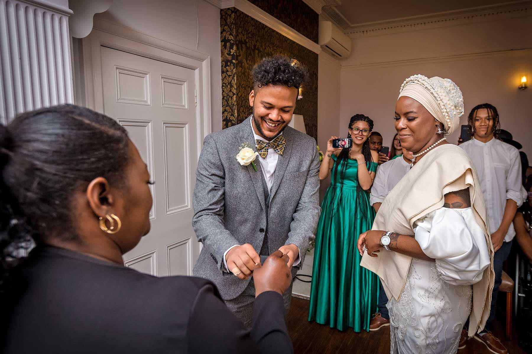 The smiling groom takes his ring from the registrar as his bride looks on in the Garden Room, Southwark Register Office