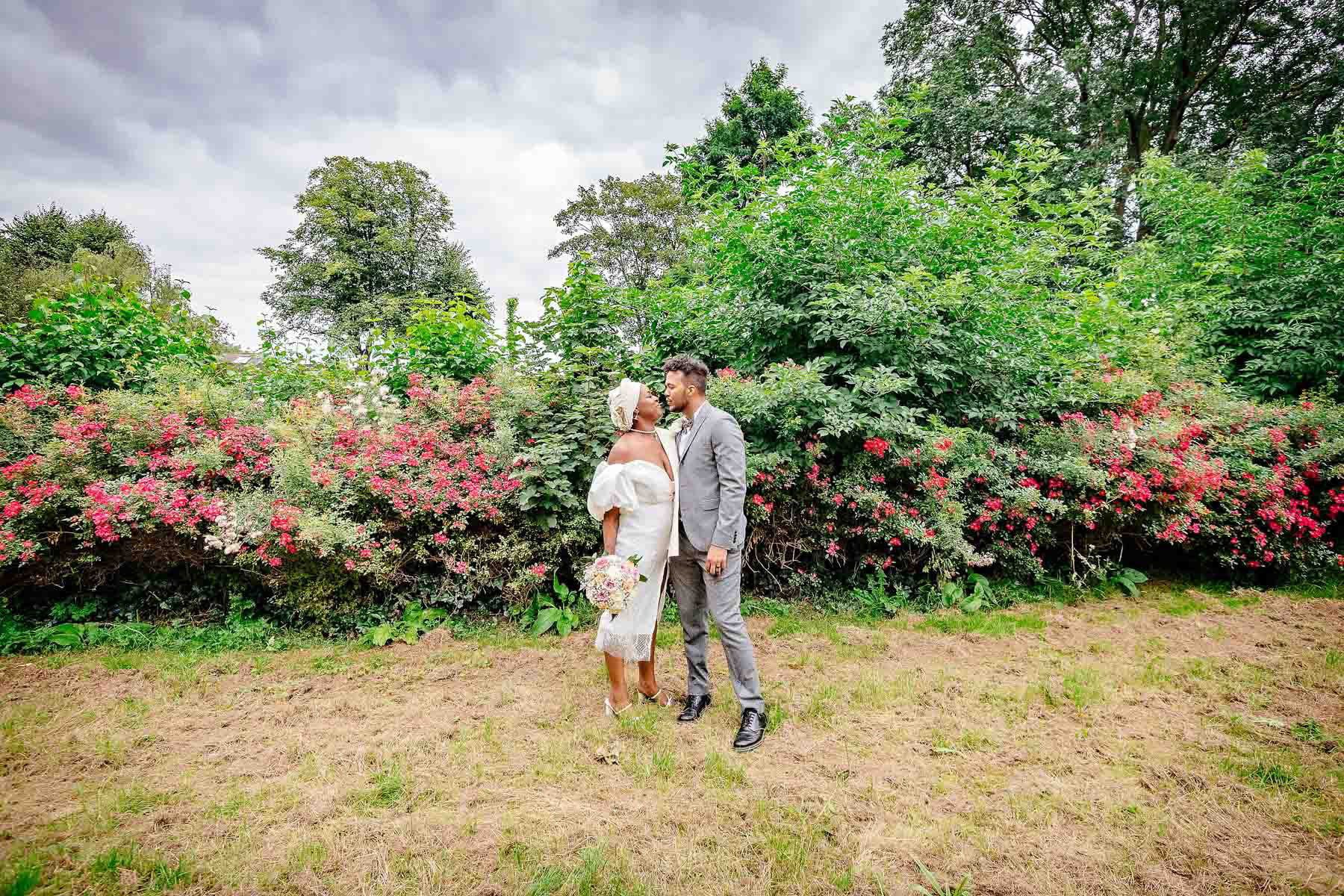 Newlyweds facing each other with red flowered bushes in the background in Lucas Gardens, Southwark