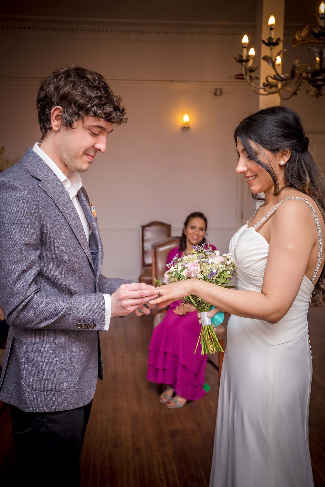The groom places the wedding ring on his smiling bride's finger in The Garen Room at Southwark