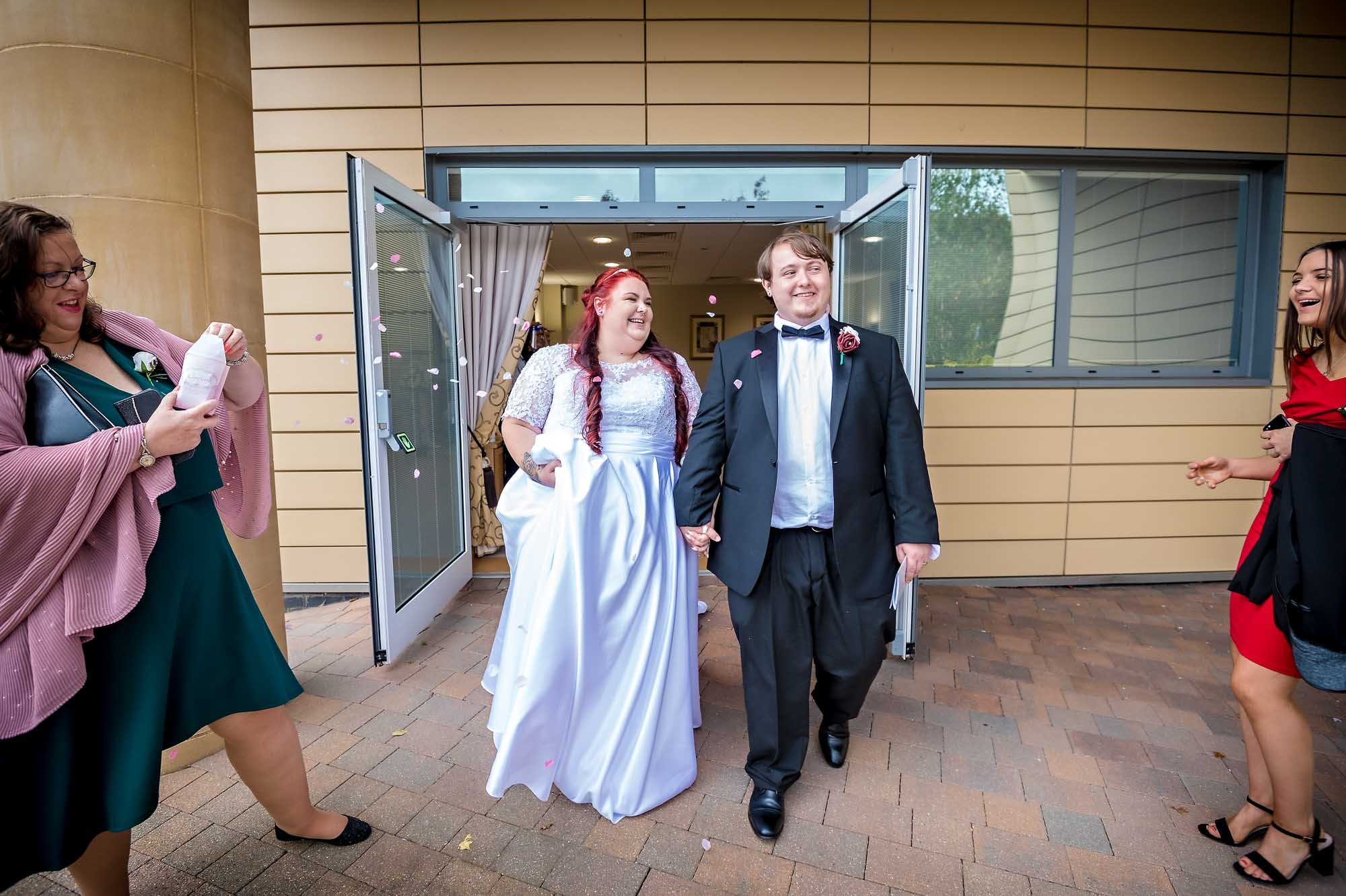 The newly married couple leave the Ceremony Room with 2 guests throwing confetti