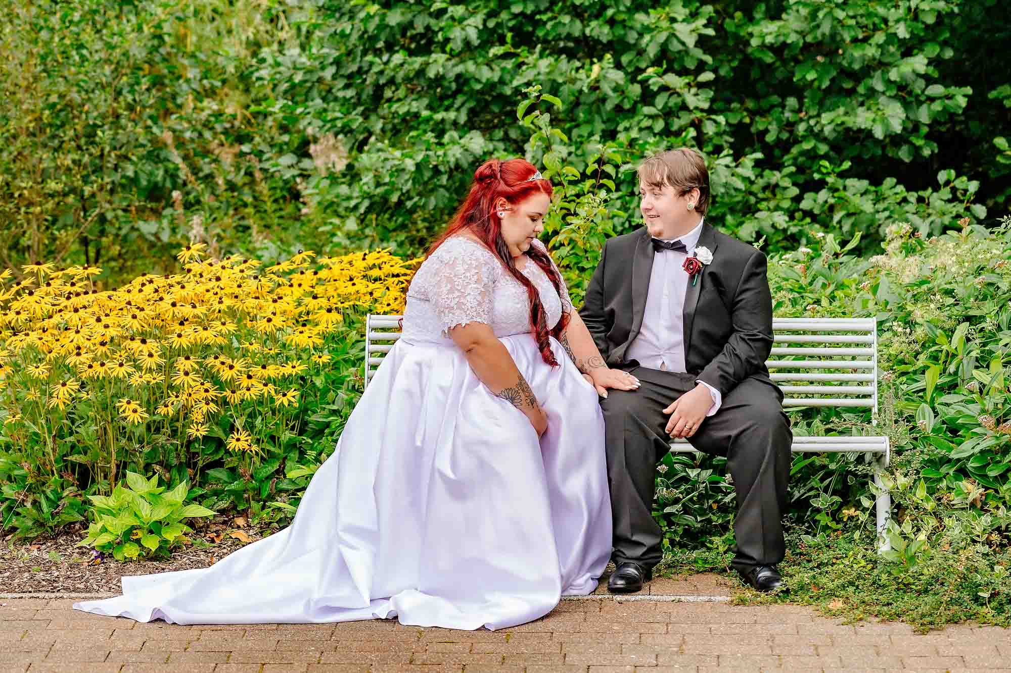Bride looking down at wedding ring with groom watching her on bench in garden of Penallta House
