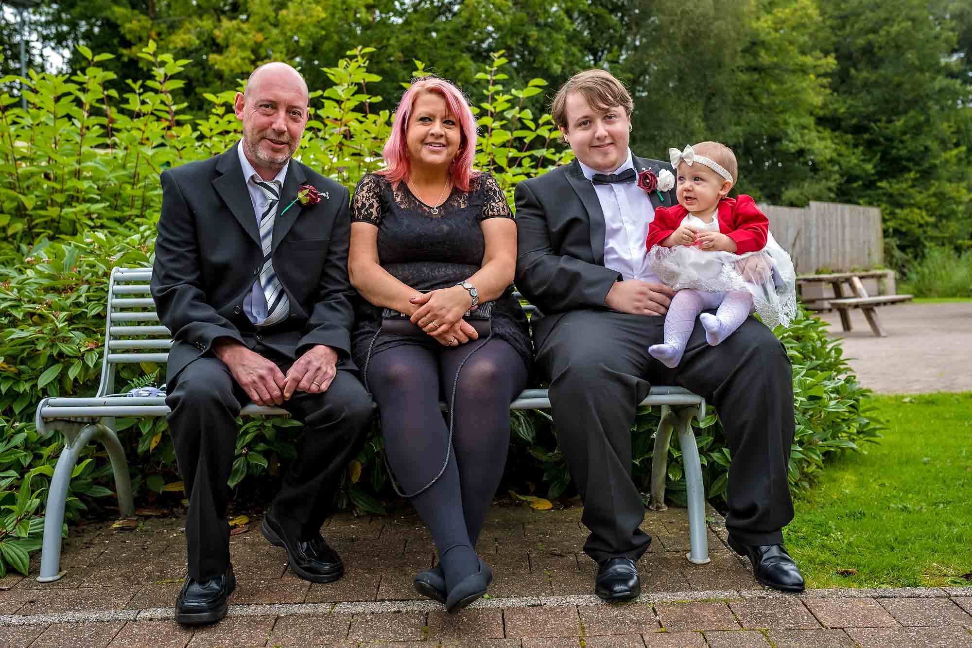 Posed wedding photo of groom with baby daughter and two guests on bench