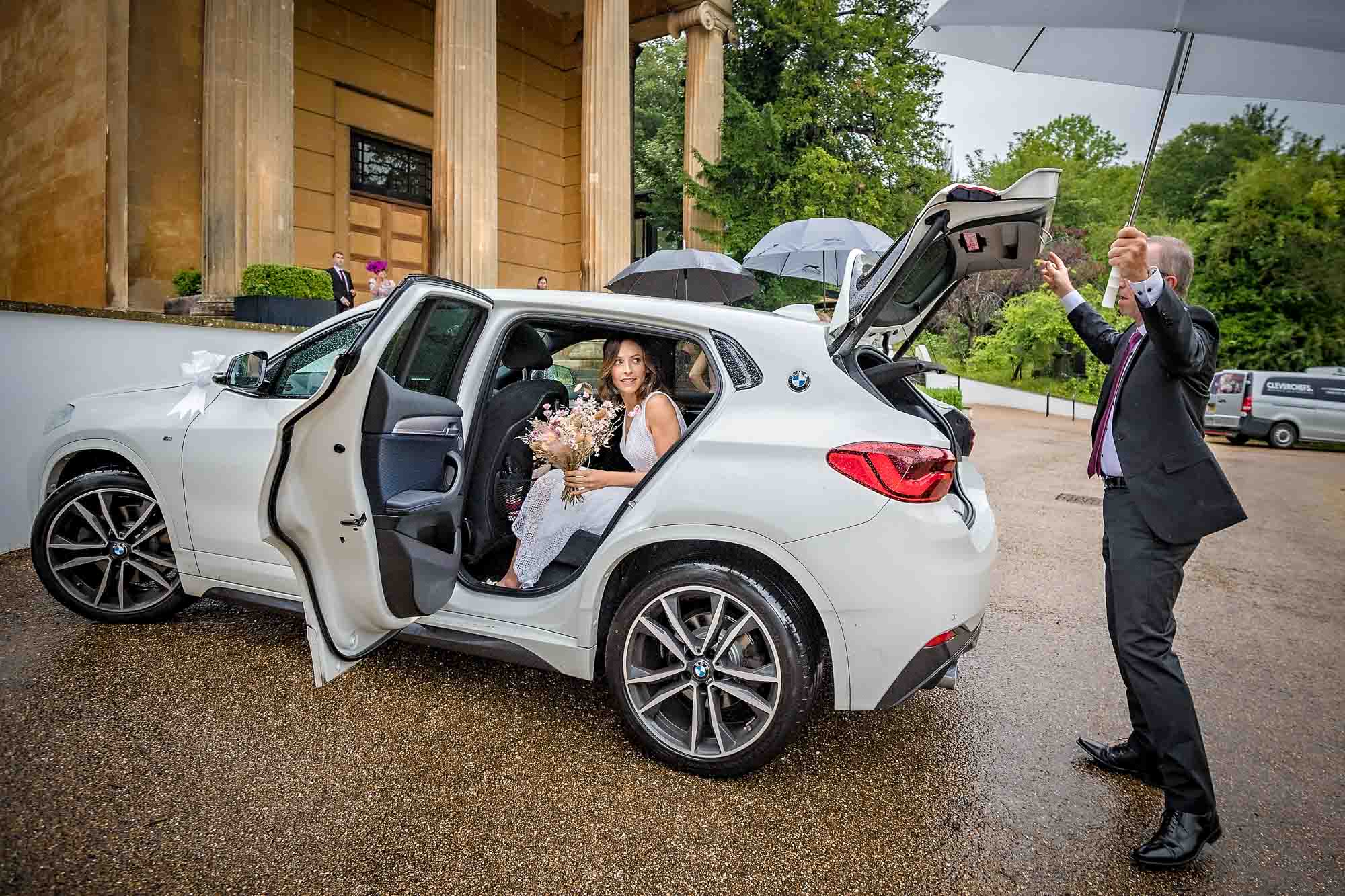 Bride looking out of car door at father at boot with an umbrella