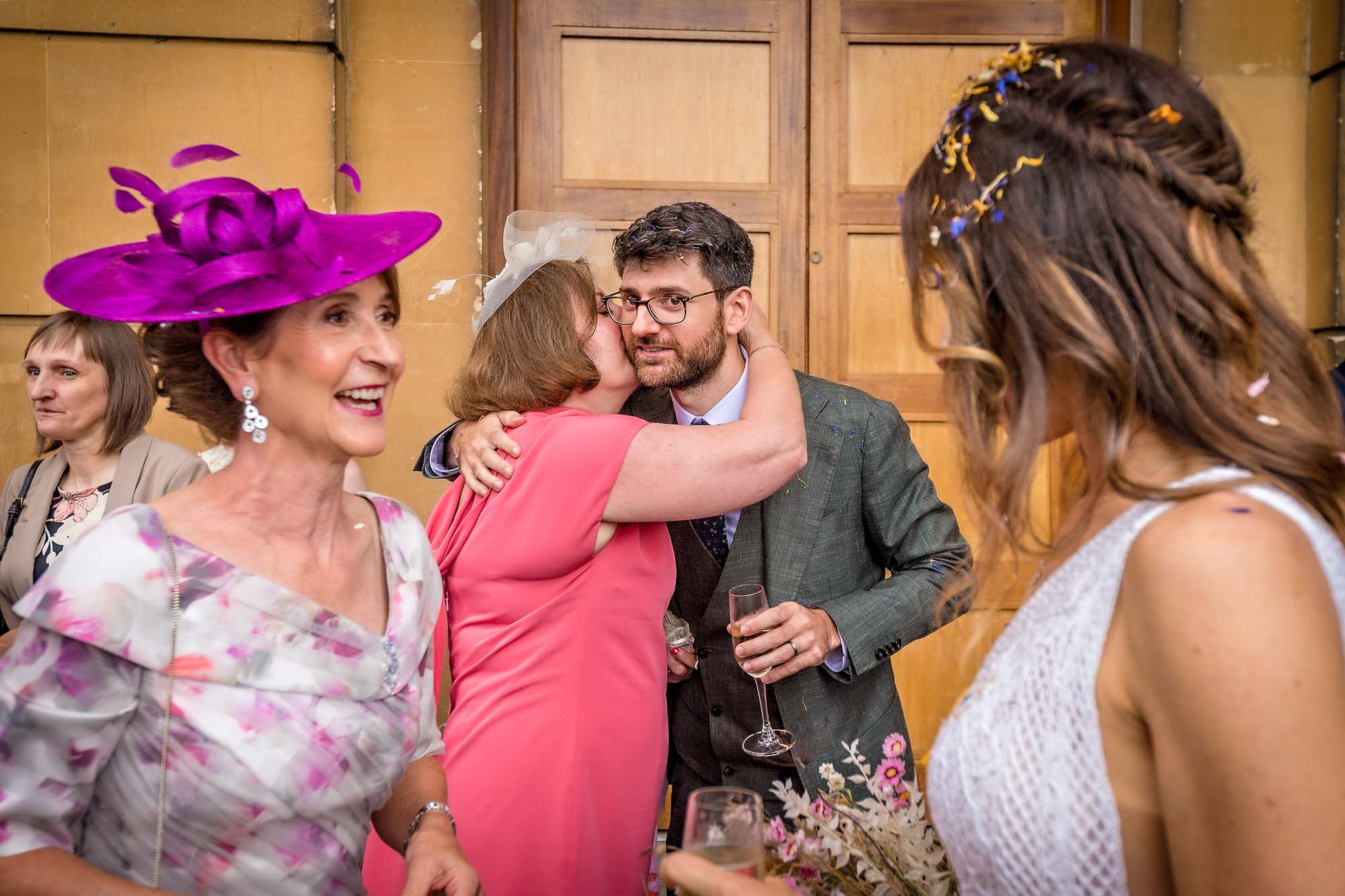 The groom's mother hugs him as he looks into camera - bride and lady chatting in foreground