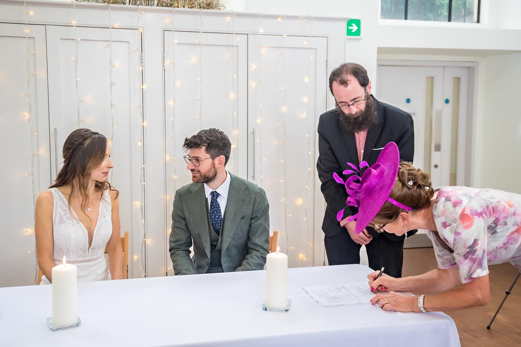 A guest signs the schedule at a wedding whilst the seated bride and groom chat to each other