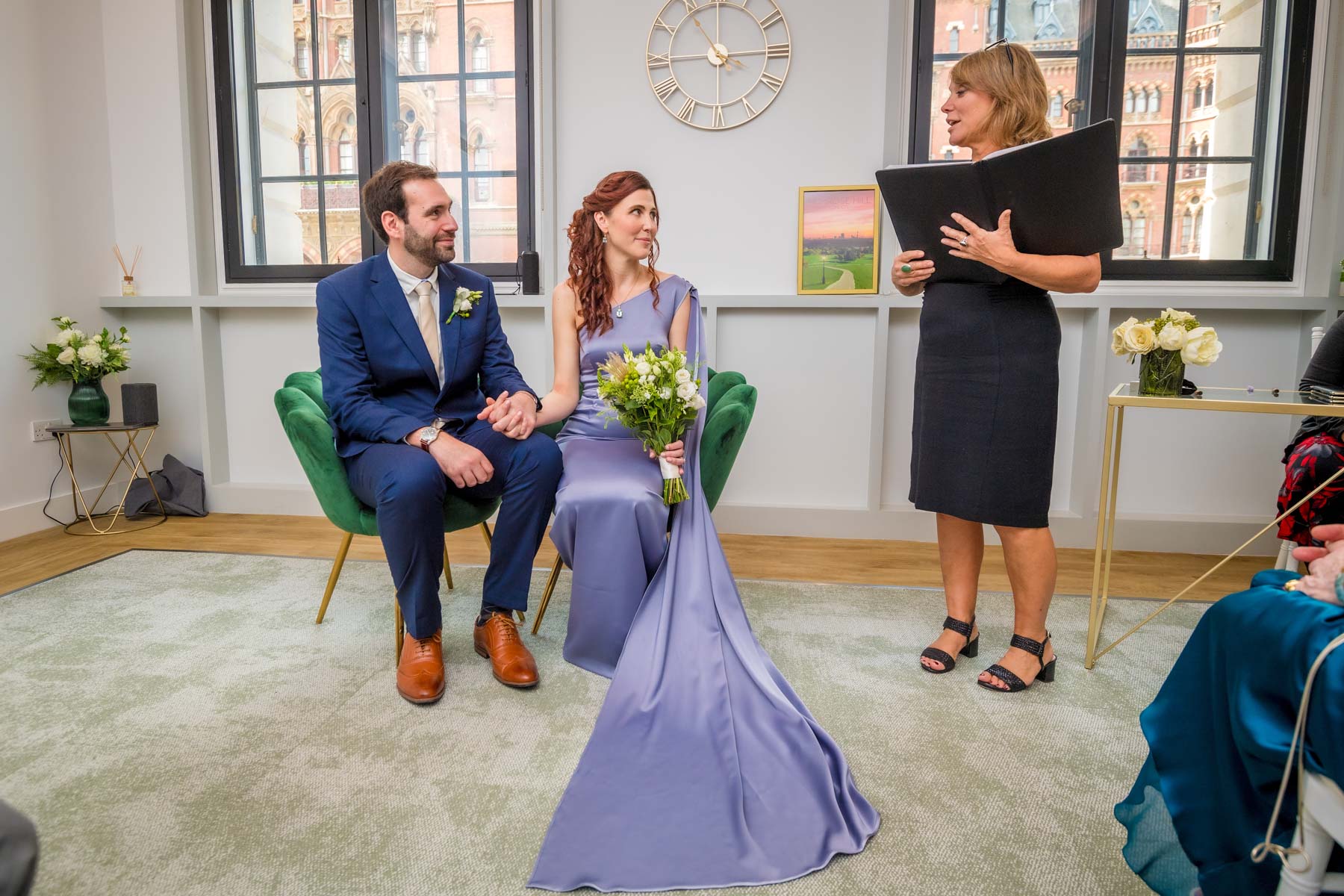 The registrar begins a wedding ceremony standing with the seated couple in Camden, London