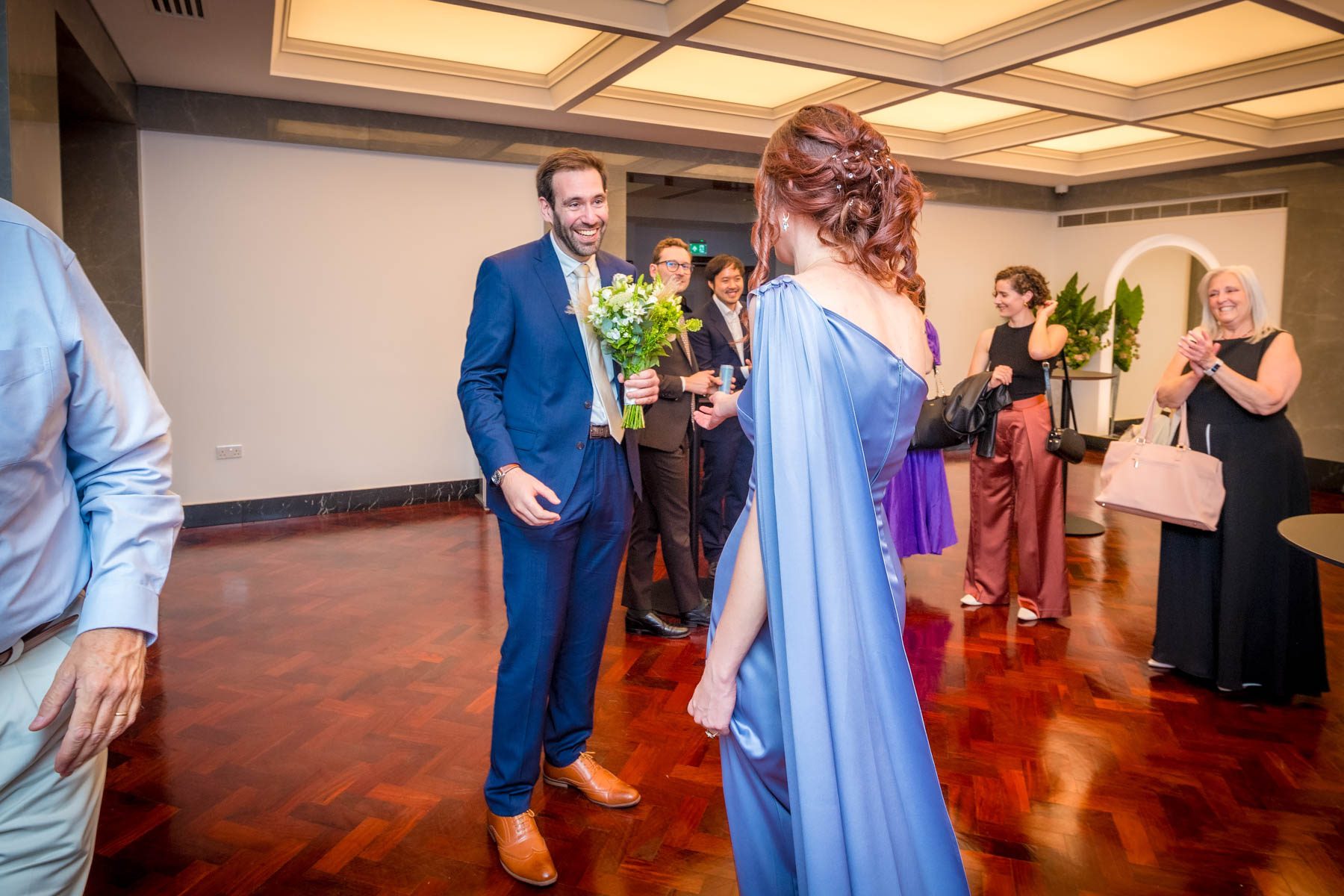 The groom smiles broadly as he sees his bride for the first time. Guests looking on.