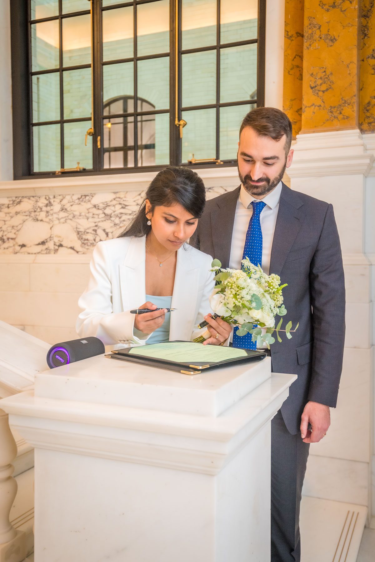 The bride holds a pen whilst signing the schedule on the Marble Staircase at Camden Town Hall