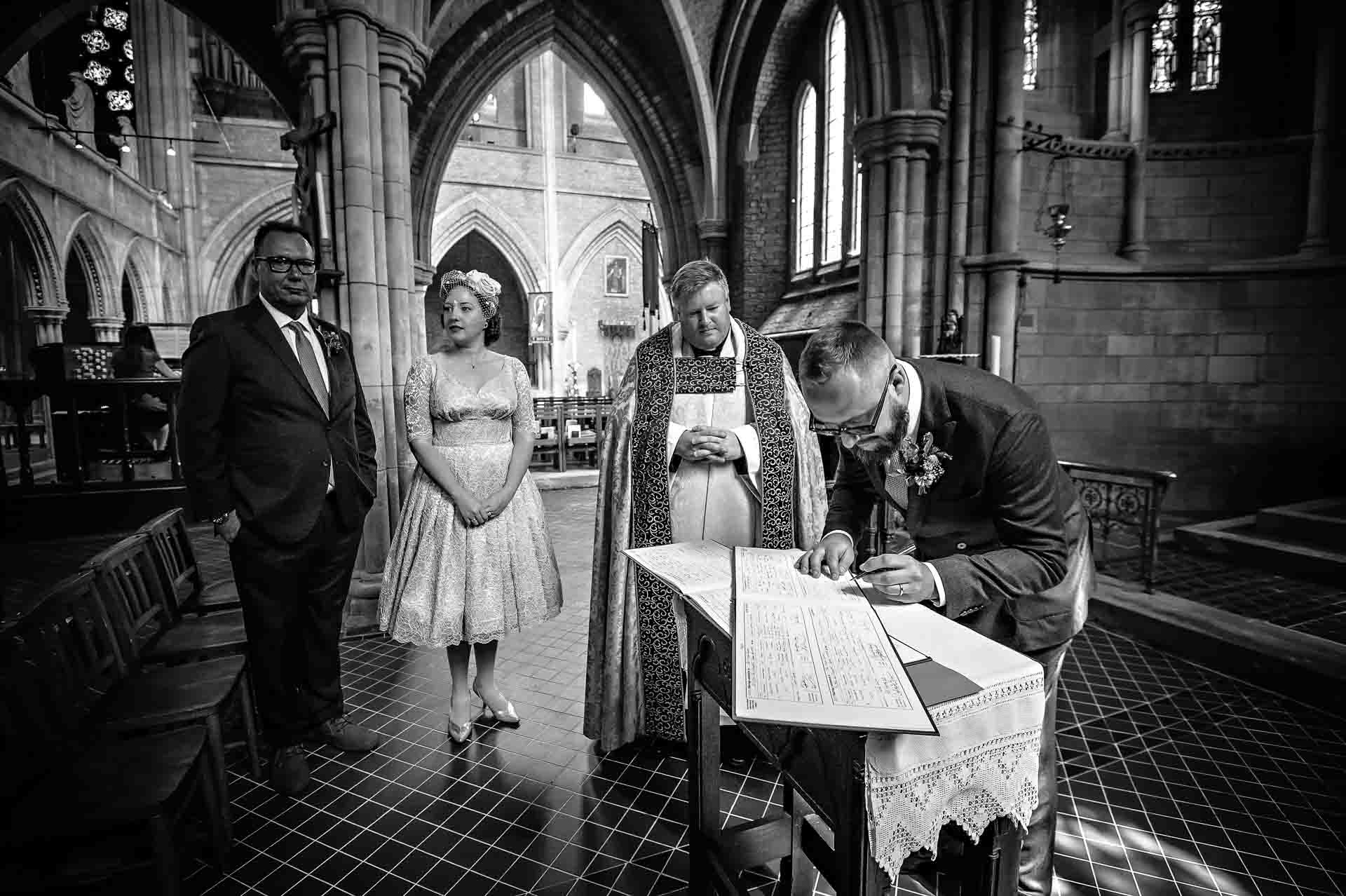 Groom signing wedding register in church wedding with vicar, bride and guest watching