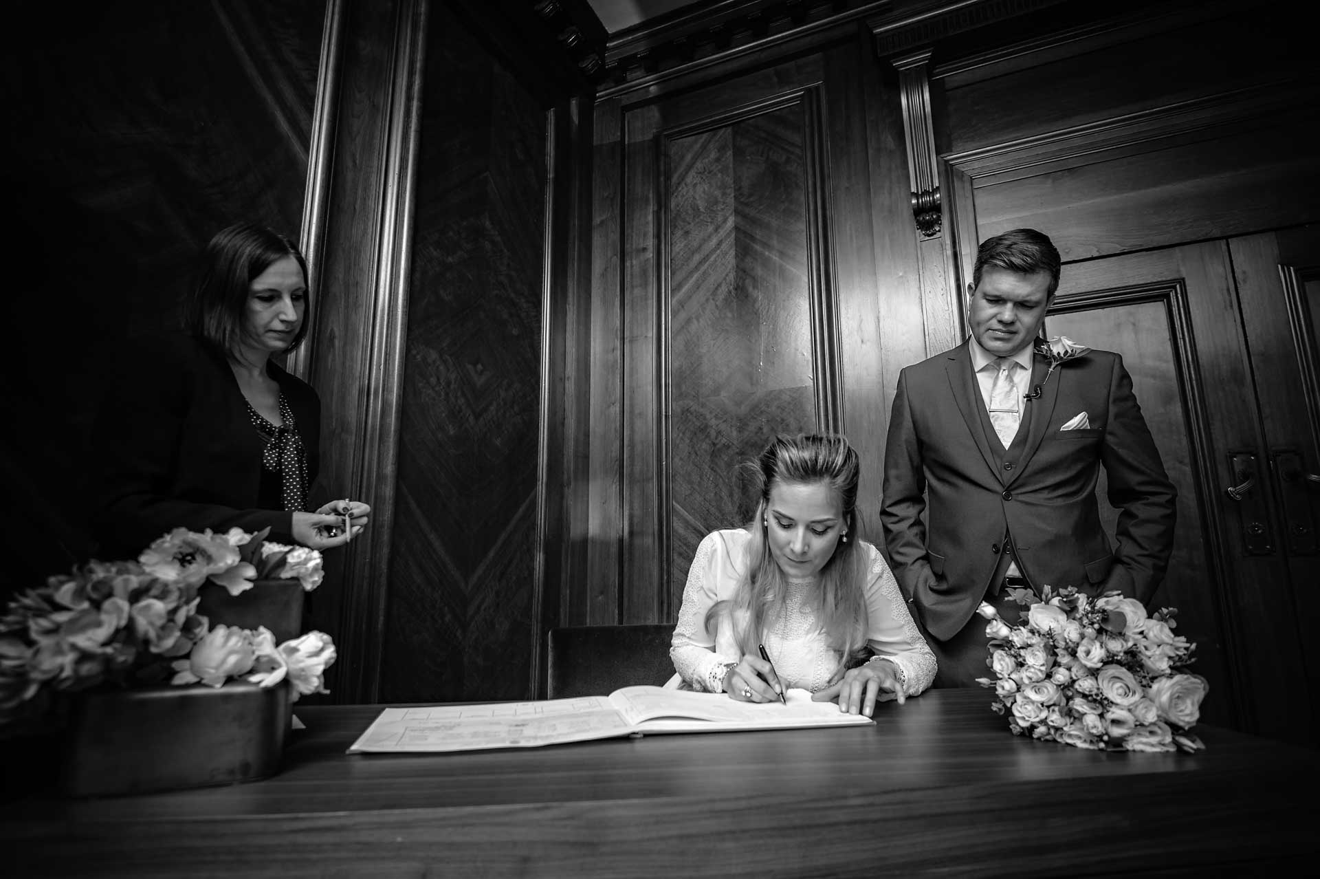 Bride signing register with groom looking on
