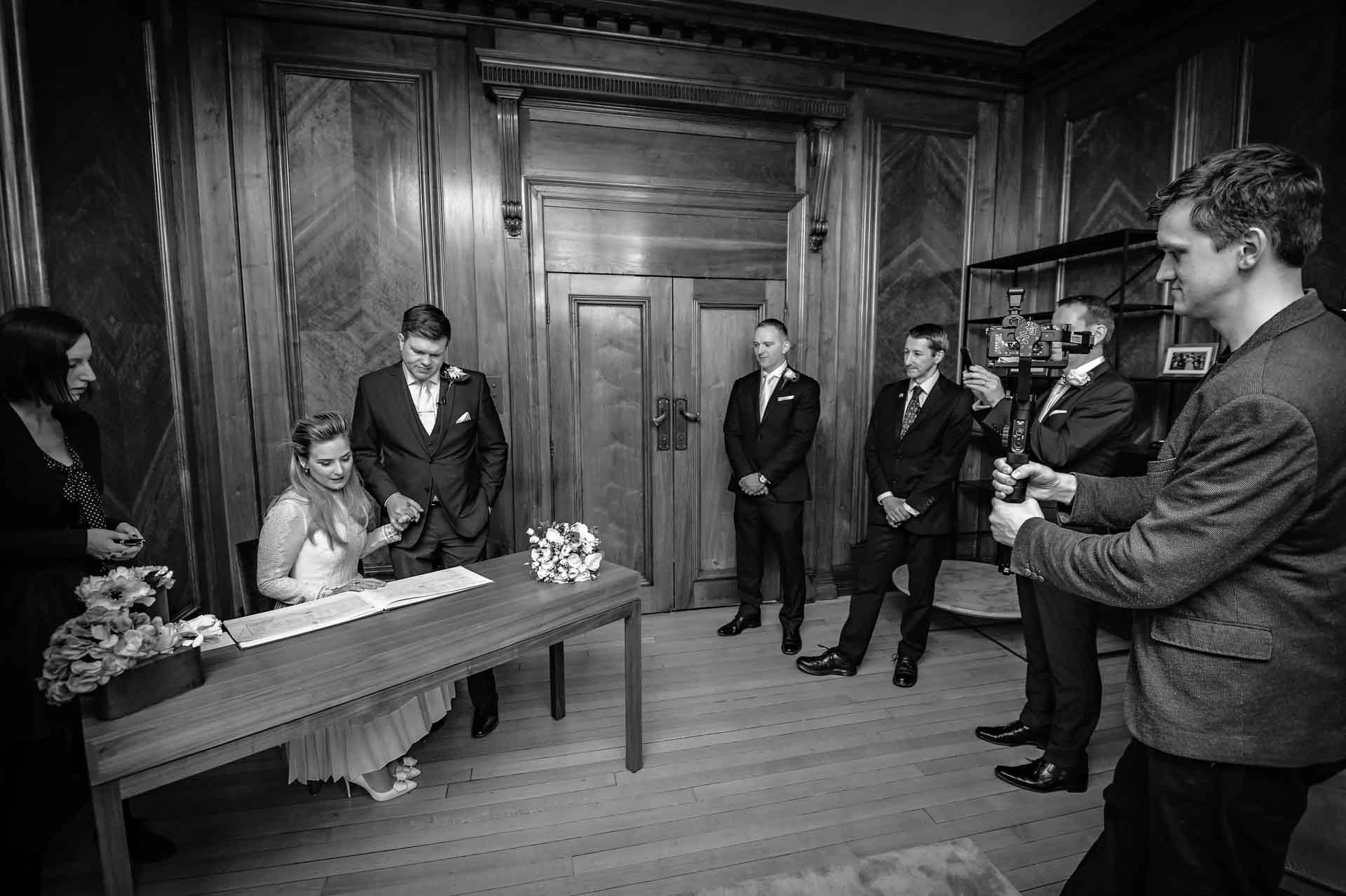 Couple at Westminster Register Office Looking at Register Whilst Guests and Videographer Watch